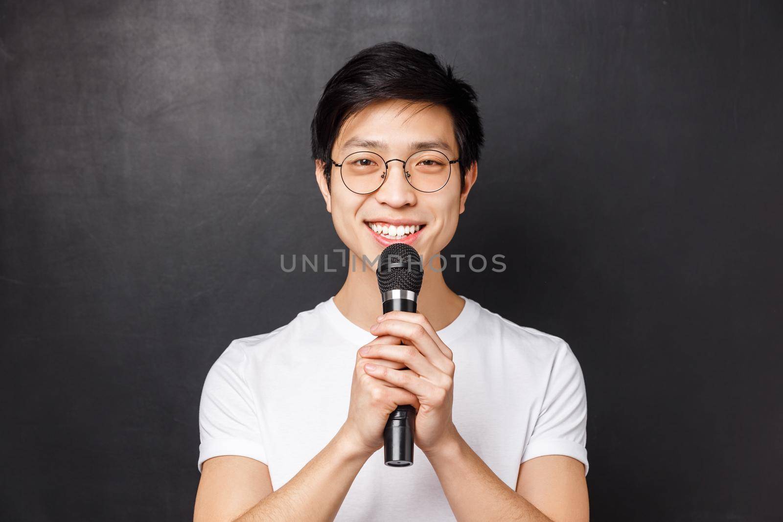 Leisure, people and music concept. Portrait of cute smiling asian man in white t-shirt, holding microphone both hands, singing song on karaoke party, perform in front of audience, black background by Benzoix