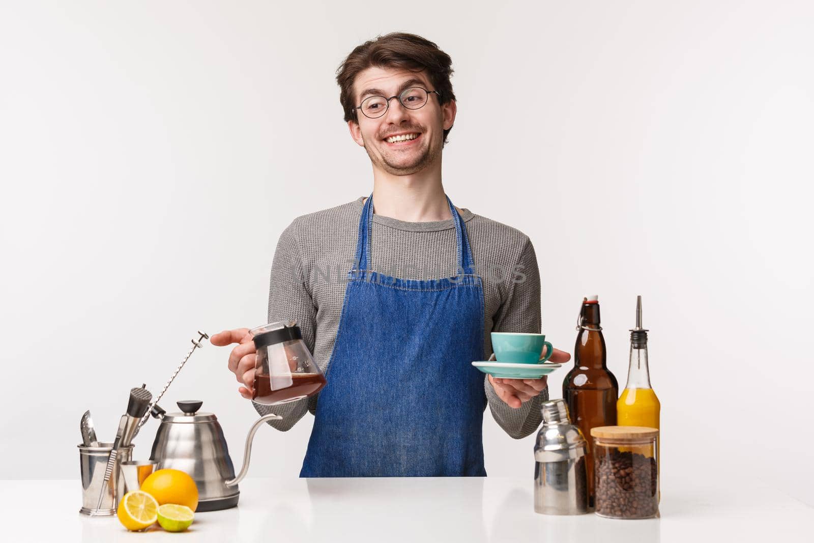 Barista, cafe worker and bartender concept. Portrait of cheerful carefree smiling caucasian man in apron, hold kettle with filter coffee and cup, laughing and look away stand bar counter.