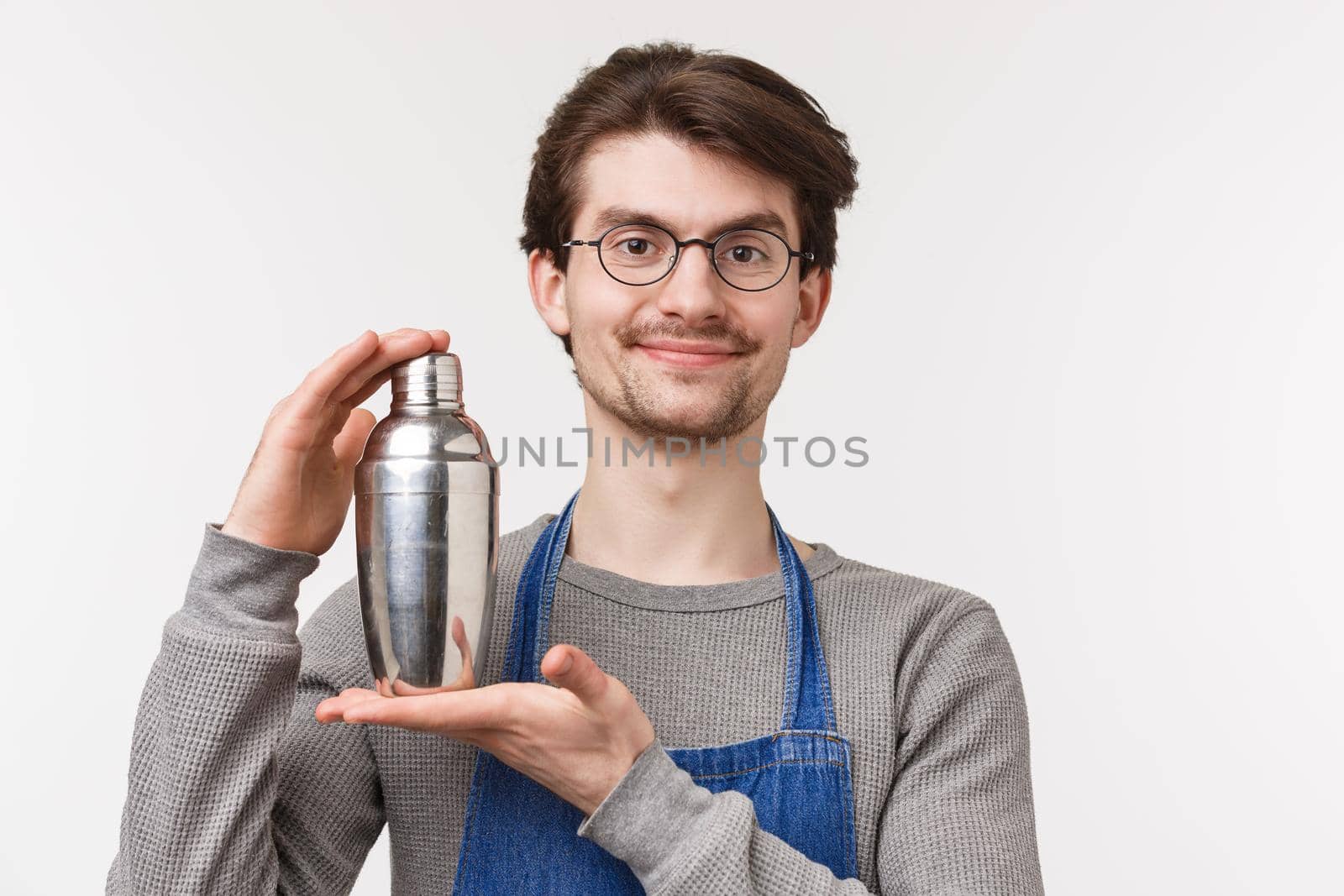Close-up portrait of cheerful smiling young bartender working on inventing new cocktail, shaking drink in shaker and smiling camera, prepare drink for customer, standing white background.