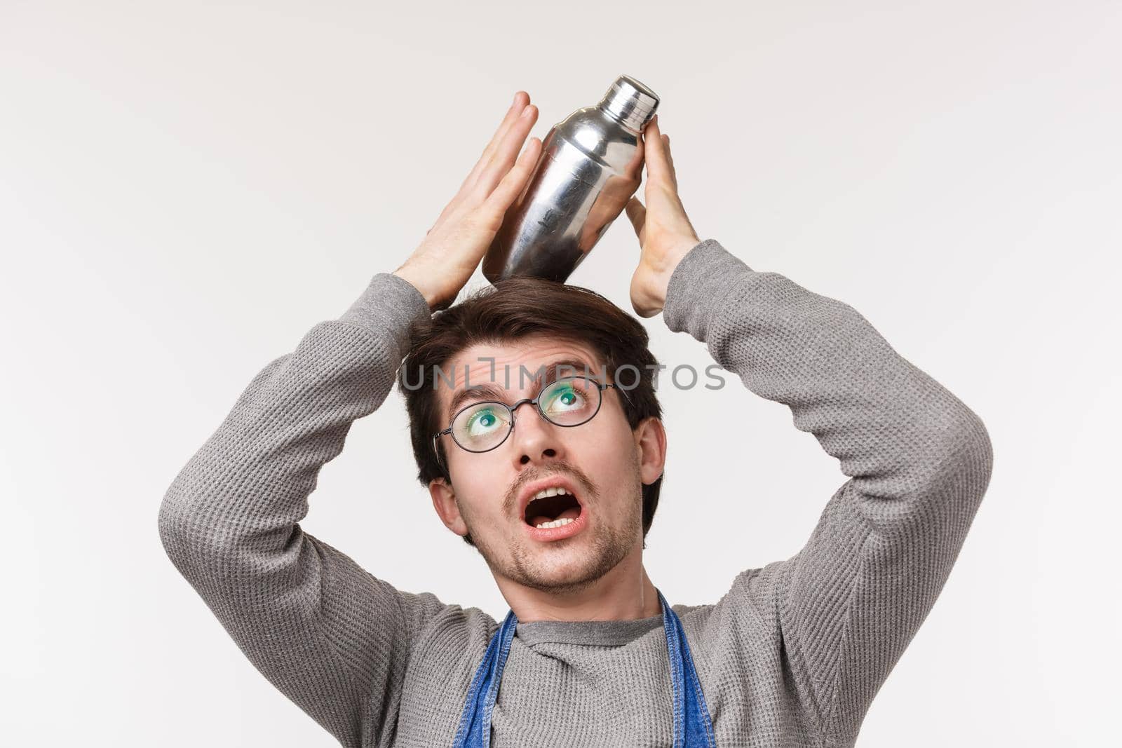Close-up portrait of funny caucasian male bartender in apron and glasses balancing shaker on head as its falling on ground, standing white background fool around while there are no customers.