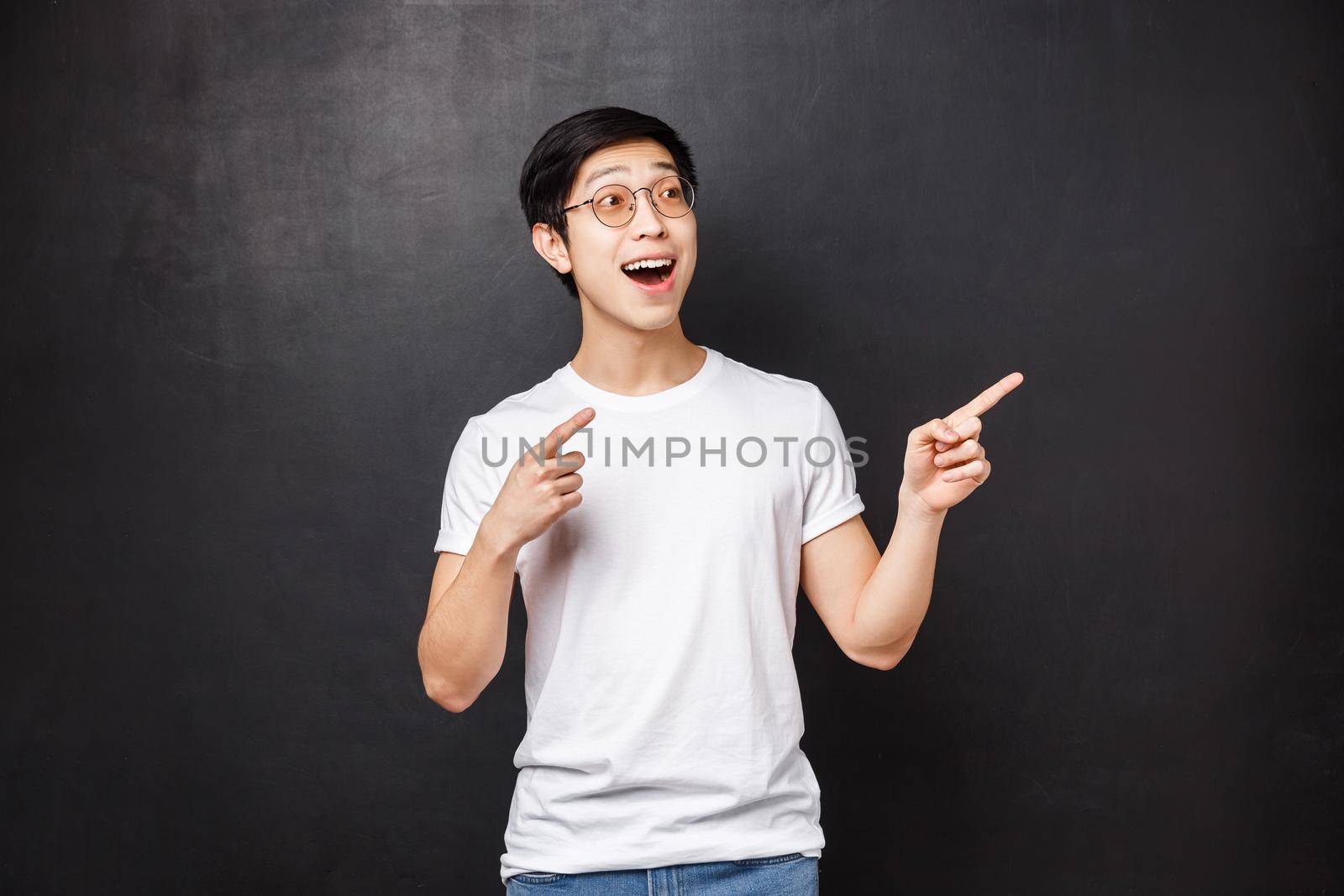 Excited good-looking asian guy in sunglasses seeing cool performance at anime festival, staring amused and thrilled pointing fingers right, express happiness and enthusiasm, black background.