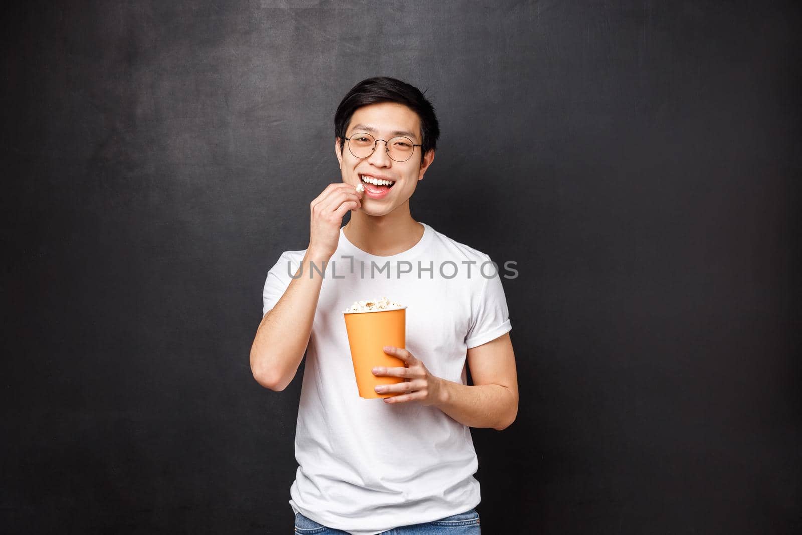 Portrait of carefree good-looking asian male attend cinema, pleased smiling and eating popcorn, watching interesting movie, enjoying weekends alone with horror, black background.