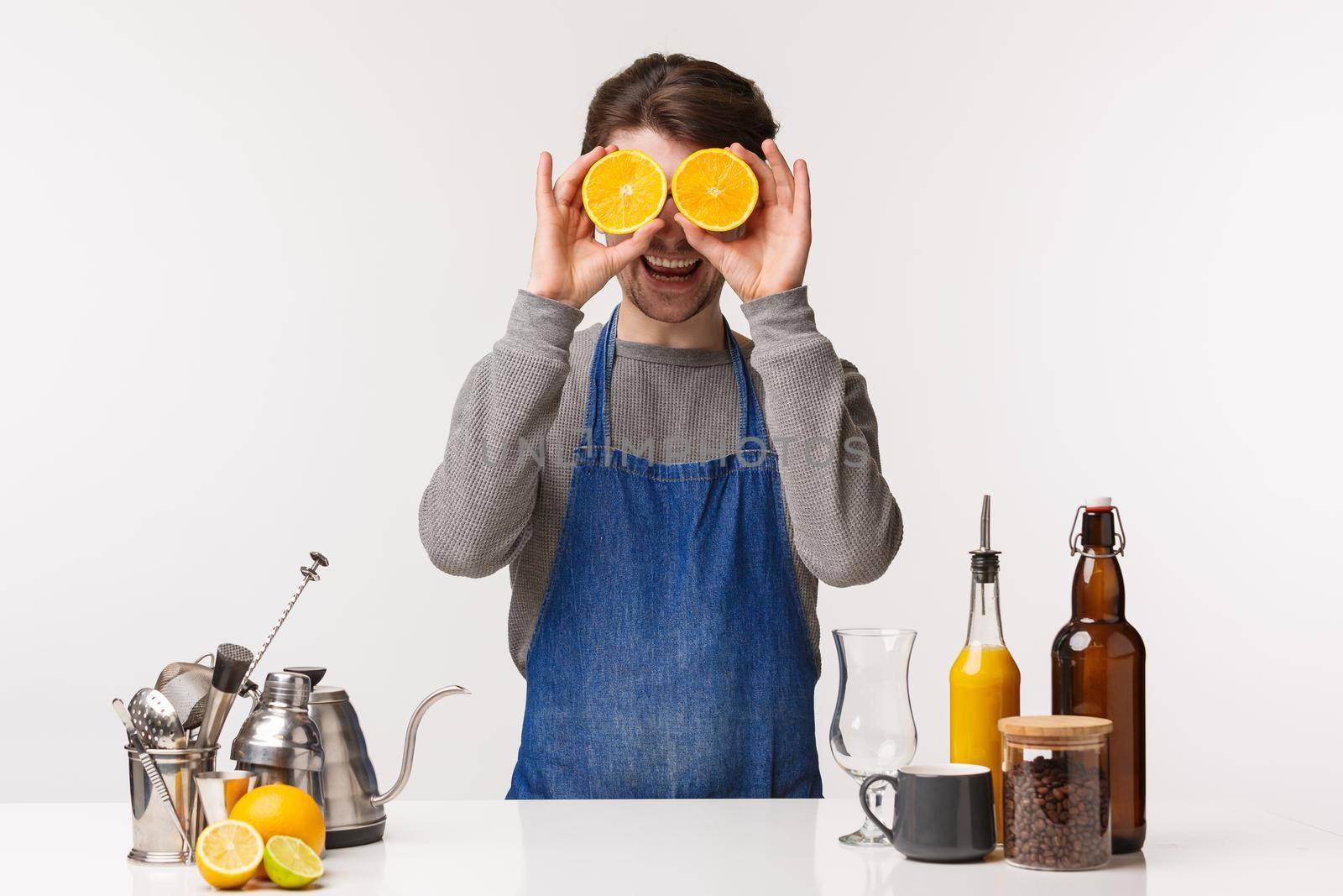 Barista, cafe worker and bartender concept. Portrait of cheerful happy male employee in blue apron making drink, holding two slices of orange like eyes mask and smiling camera, stand near bar counter.