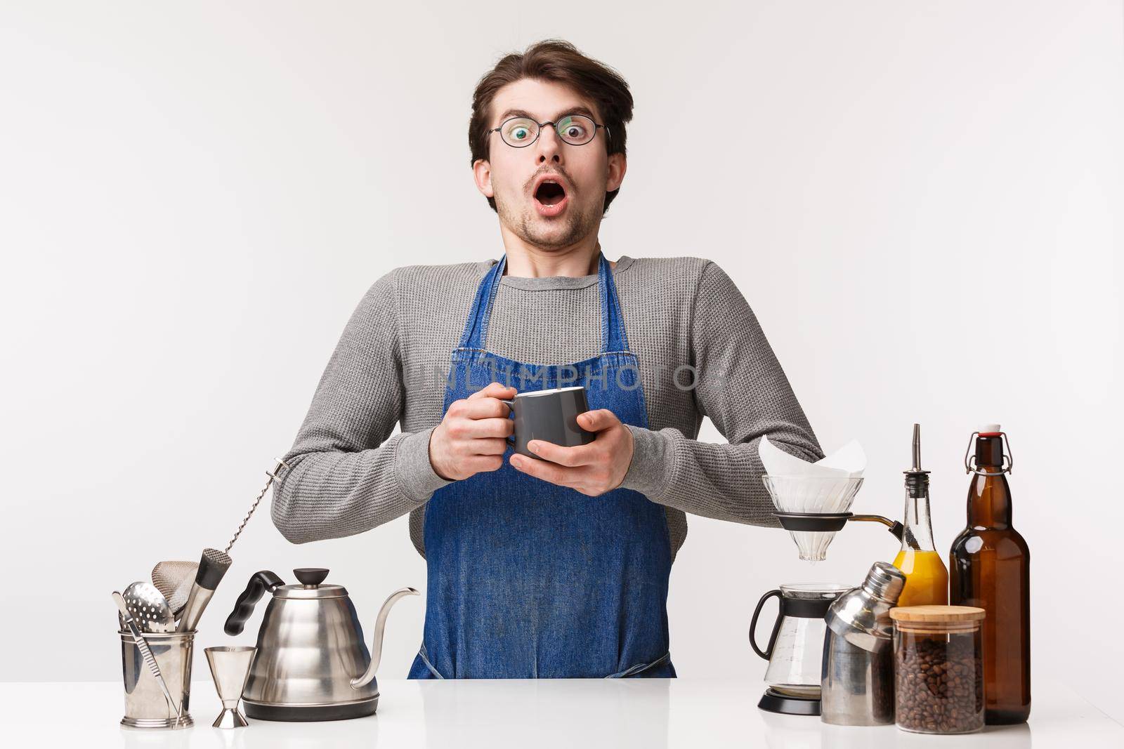 Barista, cafe worker and bartender concept. Shocked startled young male employee in apron, gasping astonished holding tea cup and staring amazed being scared while prepare coffee, white background.