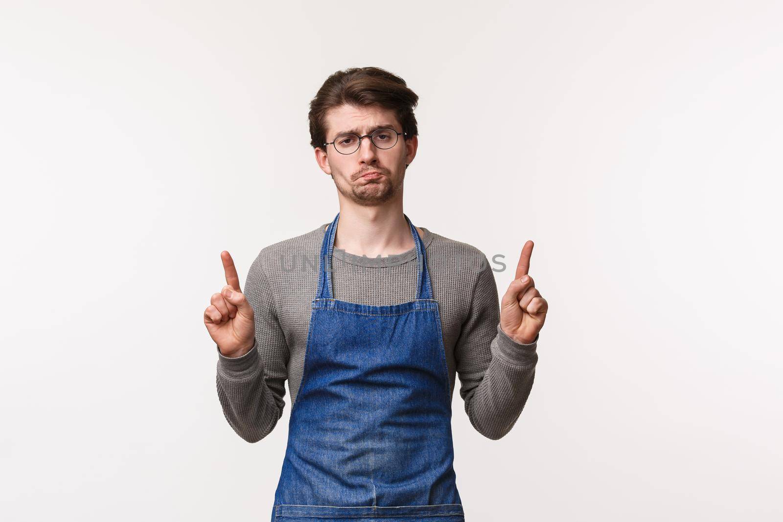 Portrait of gloomy and indifferent young bored guy working coffee shop or restaurant over bar counter, wear apron, grimace bothered and upset as pointing up at something disappointing.