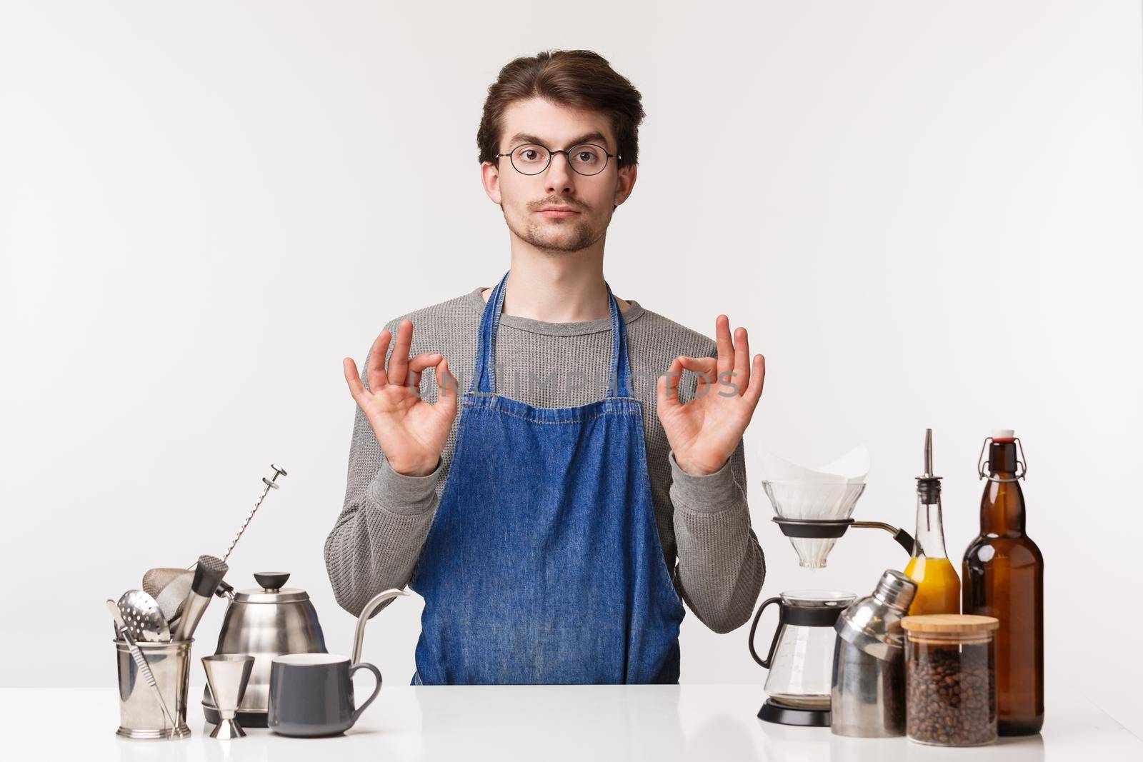 Barista, cafe worker and bartender concept. Portrait of serious-looking young caucasian guy in glasses and apron, show okay excellent sign, agree or guarantee customer will like coffee.