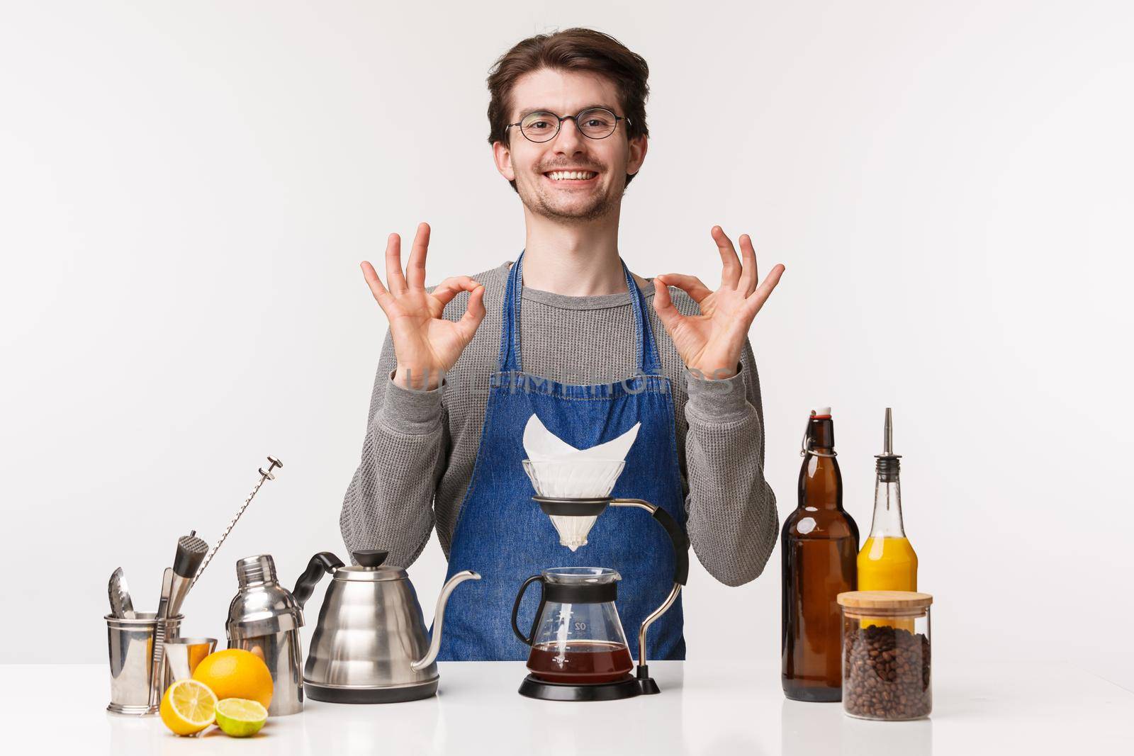 Barista, cafe worker and bartender concept. Portrait of cute friendly smiling caucasian male employee in apron guarantee you will like coffee, make filter with kettle, show ok sign.