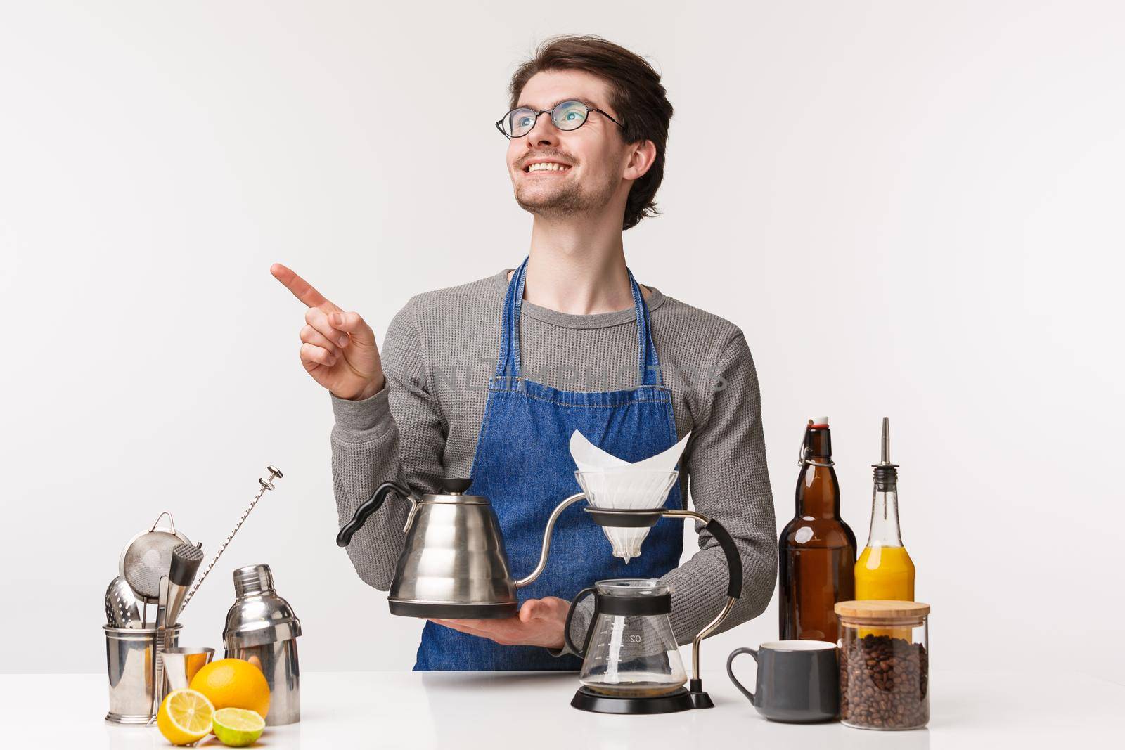 Barista, cafe worker and bartender concept. Portrait of dreamy handsome smiling bearded man in apron, holding kettle and pointing upper left corner with pleased face