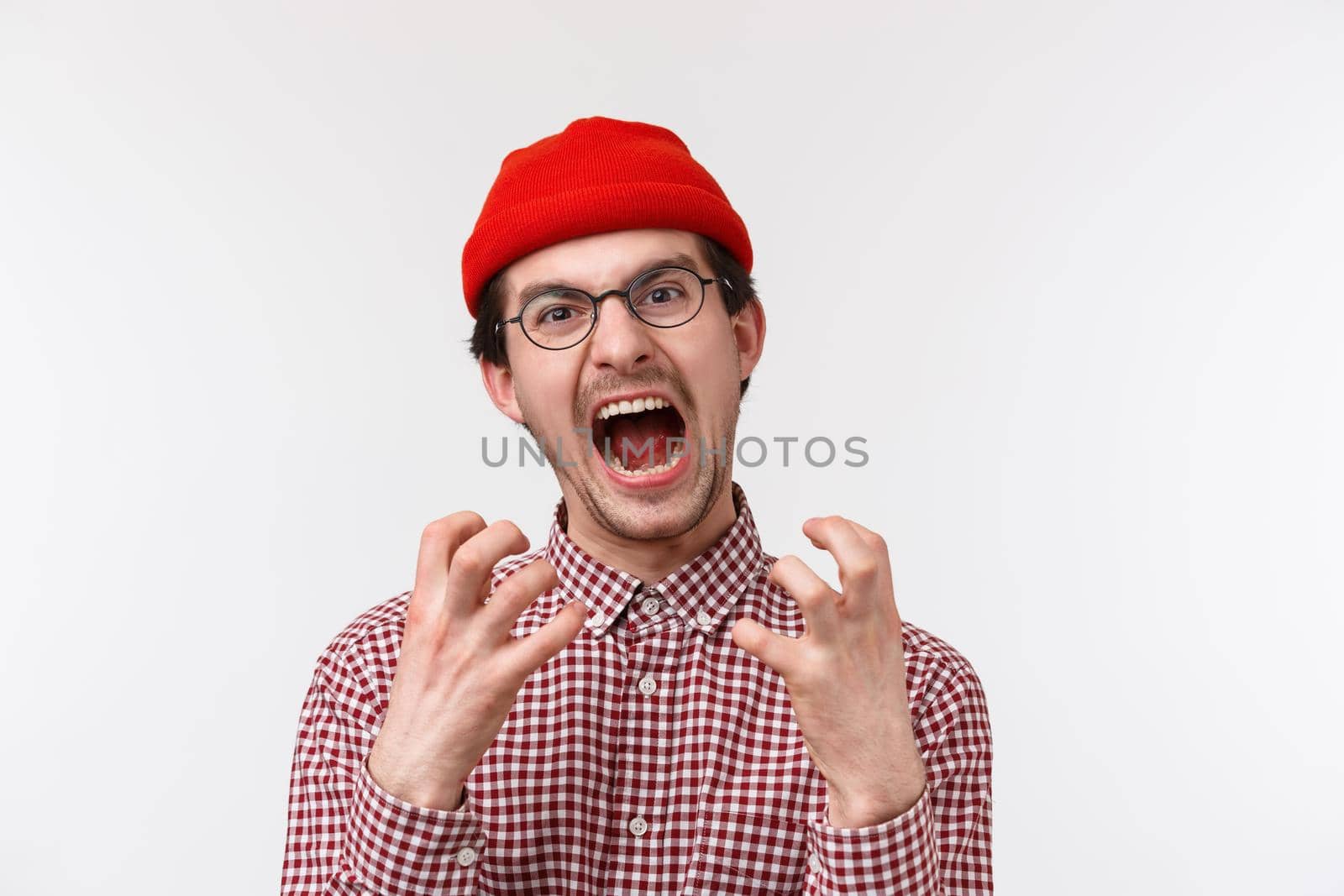 Close-up portrait of distressed and angry young funny hipster guy with moustache wear red beanie, glasses, squeez hands annoyed and bothered, screaming from pissed-off feeling, white background.