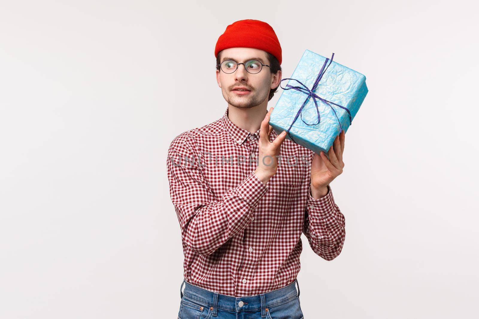 Waist-up portrait excited caucasian bearded guy in red beanie, shaking blue wrapped gift box near ear as trying guess whats inside, look thrilled, unboxing present on b-day party celebration by Benzoix