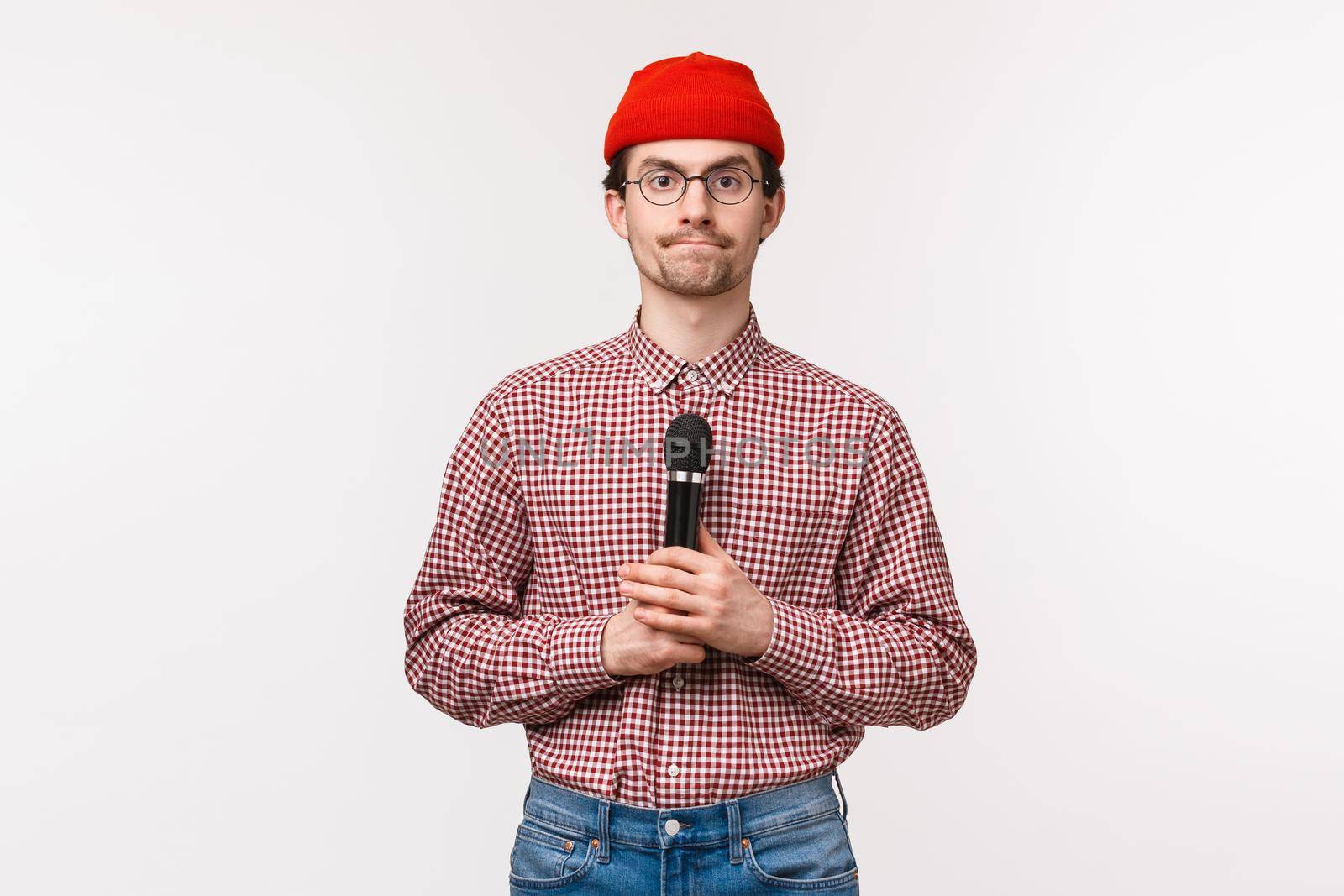 Close-up portrait of shy and geeky cute young bearded guy in glasses and red beanie, looking at tv screen and holding micrphone, getting ready to sing karaoke, standing white background by Benzoix