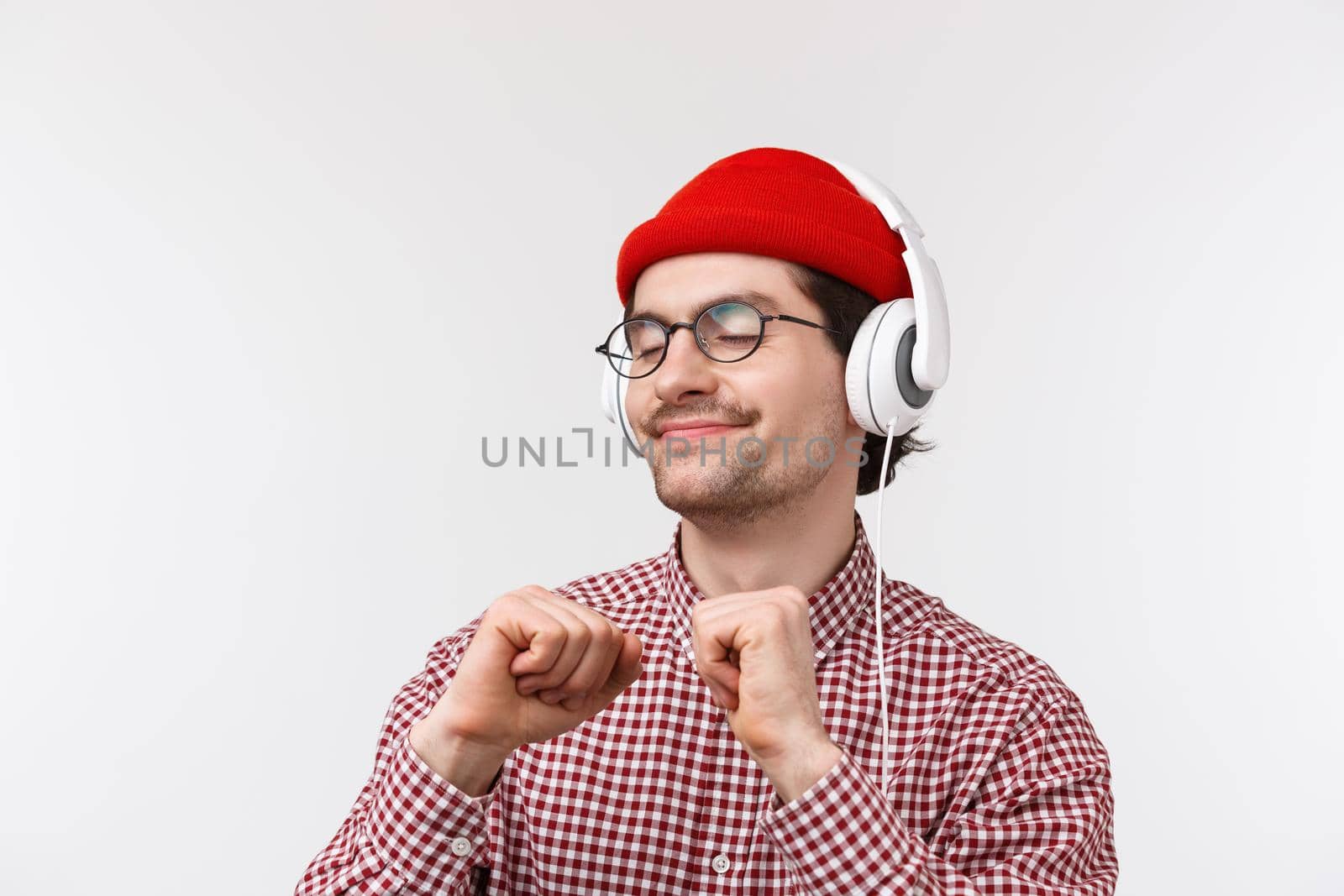 Just vibing. Happy carefree young hipster guy with moustache in glasses and red beanie dancing joyfully, listening to favorite song in headphones with pleased relaxed face, standing white background.