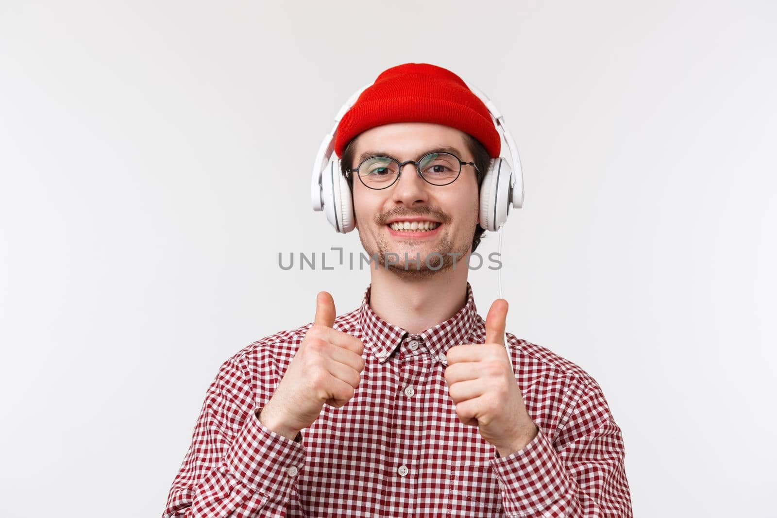 Music, technology and people concept. Close-up portrait of satisfied pleased young caucasian bearded hipster man in glasses and beanie, show thumbs-up as listening music in headphones.
