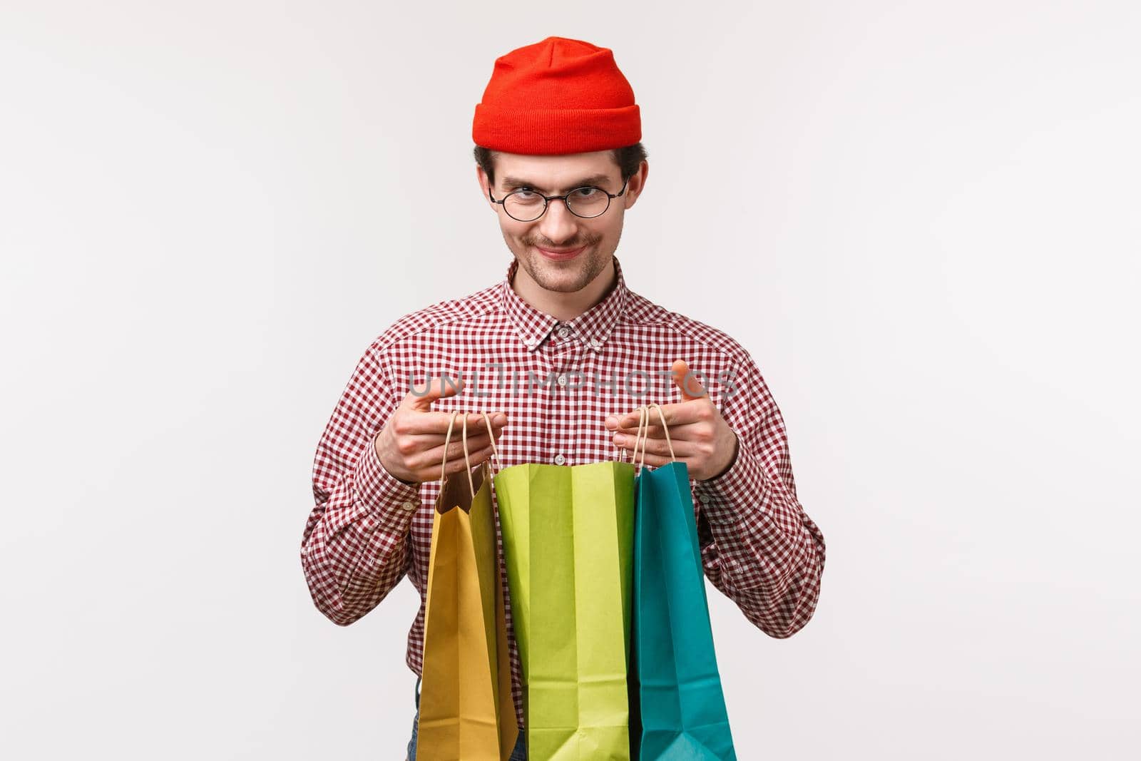 Funny and excited young caucasian hipster guy in red beanie, glasses, open shopping bags and looking at camera pleased as receive surprise present from friends, standing white background delighted.