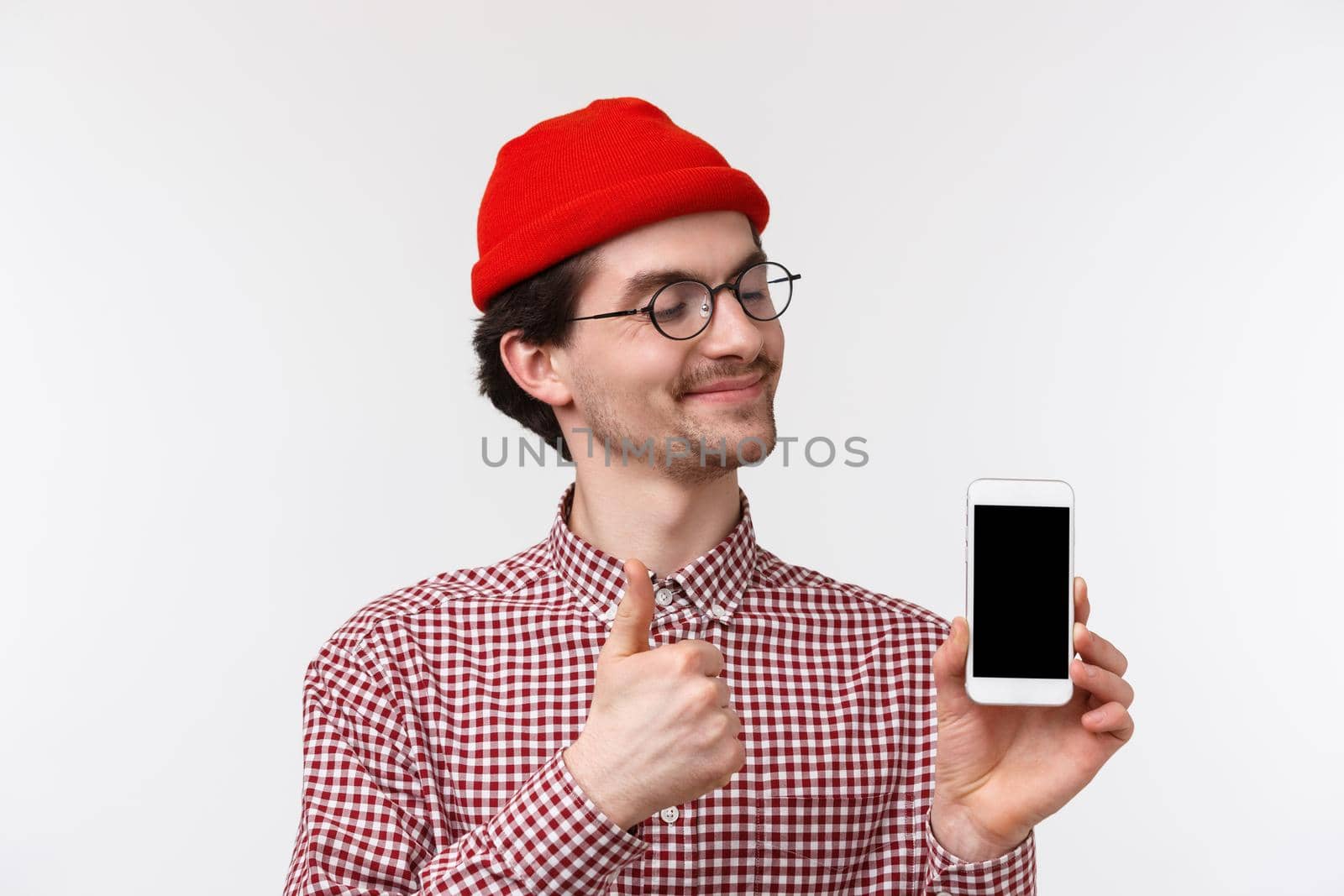 Technology and people concept. Close-up portrait pleased happy young hipster man in red beanie and glasses, looking satisfied at mobile phone display, make thumb-up approve smartphone gadget by Benzoix