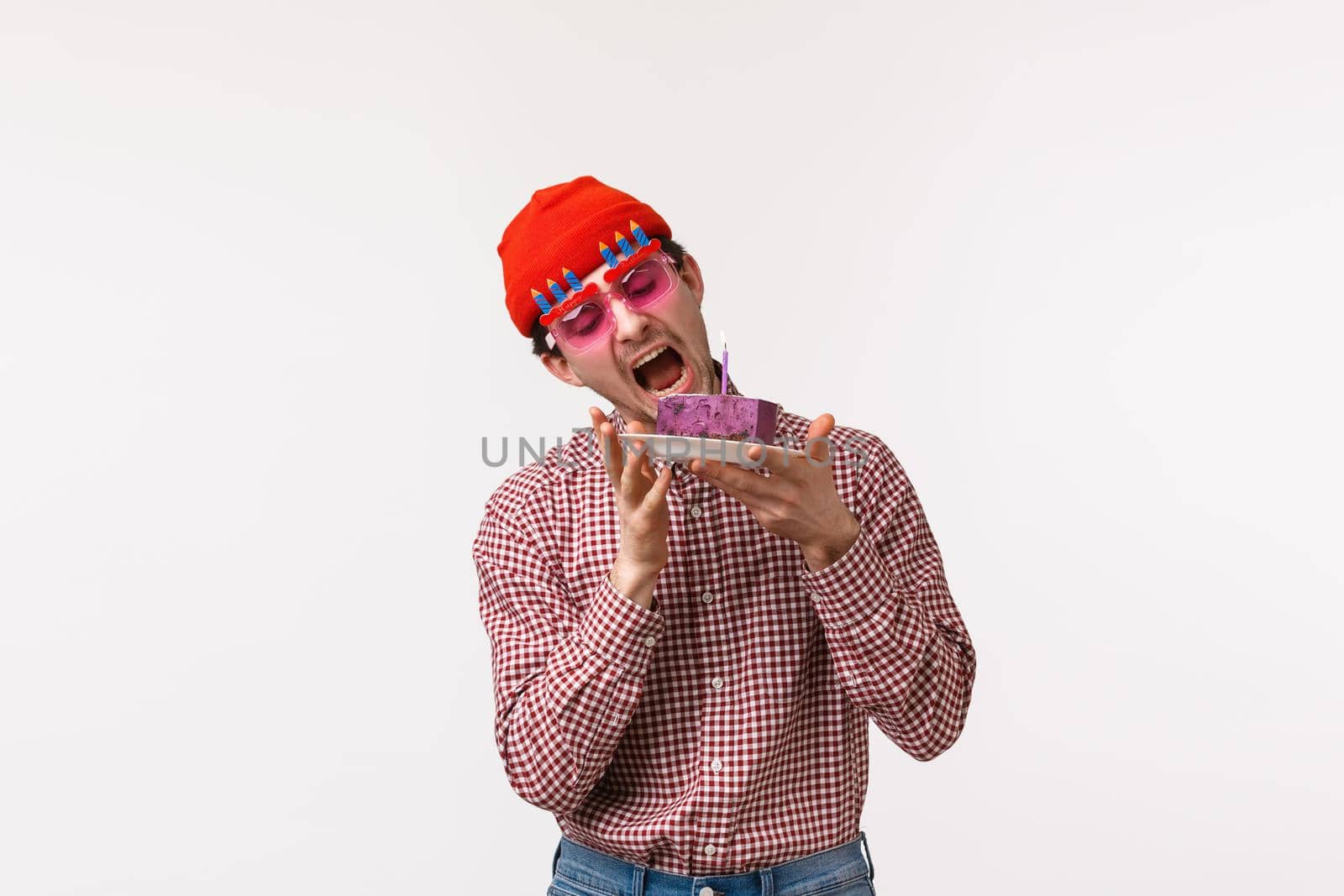 Celebration, holidays and lifestyle concept. Funny and cute adult hipster guy celebrating birthday, wear sunglasses and biting delicious b-day cake, eating tasty dessert, stand white background.