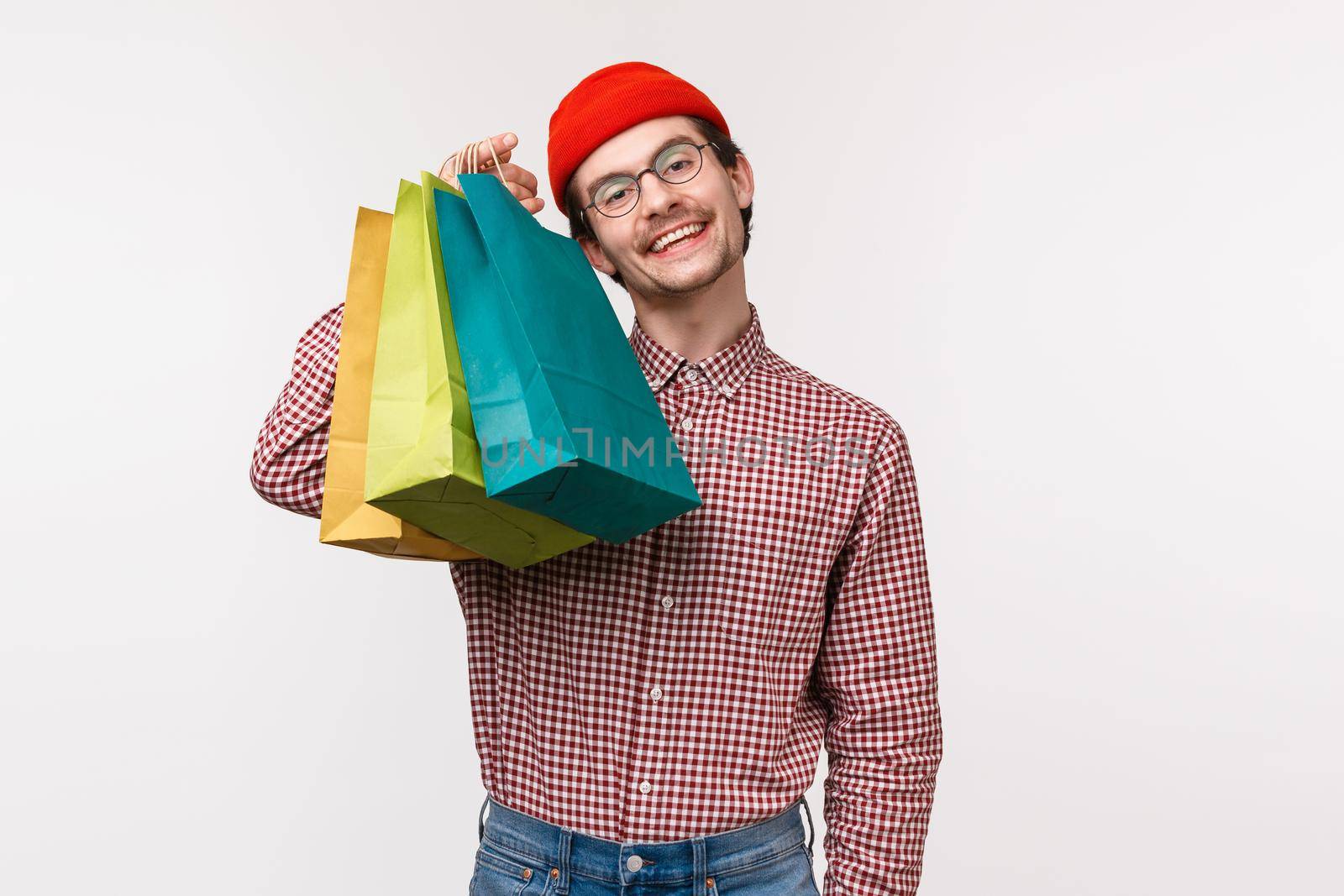 Treat yourself day. Pleased and happy smiling caucasian guy likes shopping, holding bags and look camera satisfied as bragging with his new staff he bought in store, white background by Benzoix