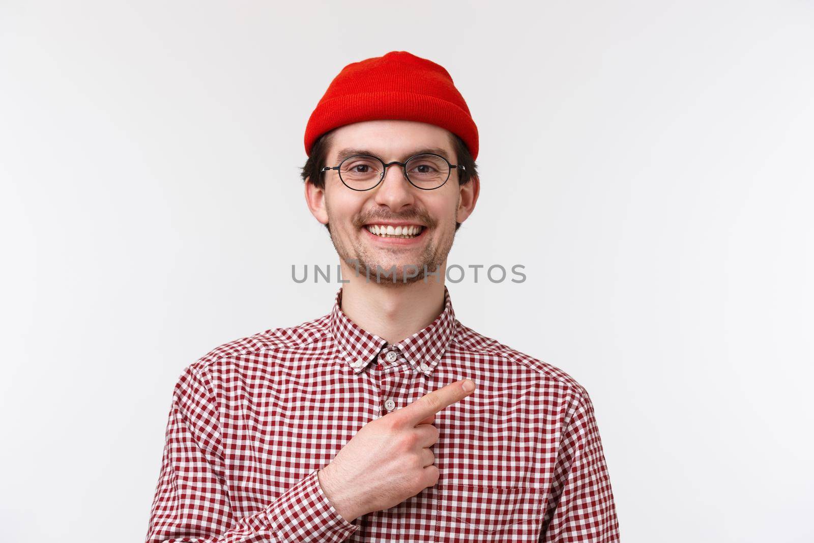 Close-up portrait handsome friendly-looking hipster male in red beanie and glasses, smiling satisfied and happy, pointing finger upper right corner, showing cool product or place hang out by Benzoix