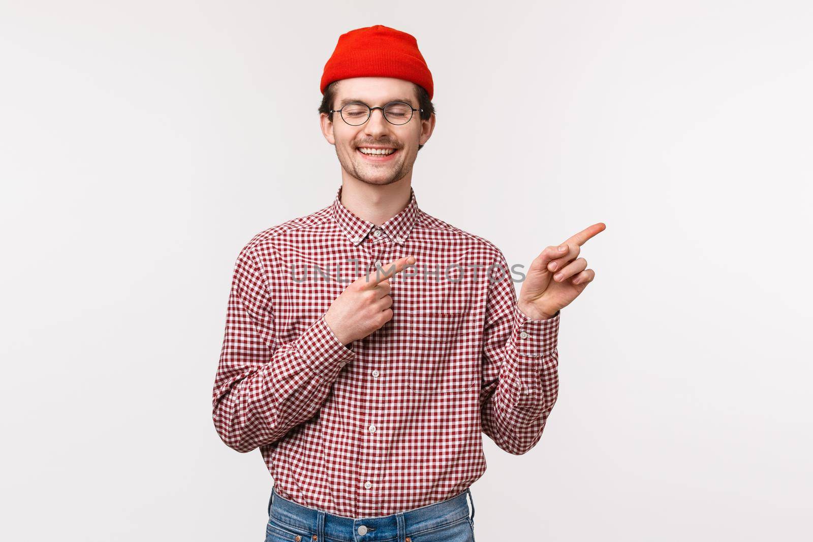 Waist-up portrait of happy, cheerful dreamy adult man with moustache, in red beanie and glasses, smiling with closed eyes as cheering found what he was looking for at upper right corner advertisement.