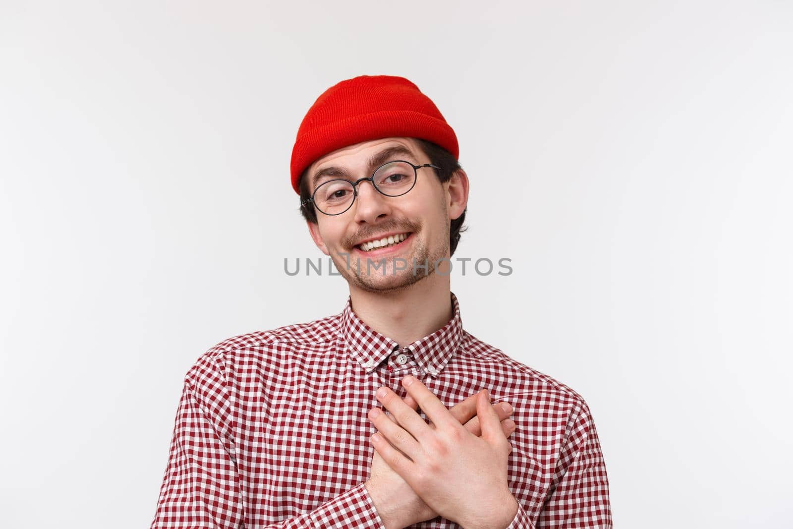 Close-up portrait funny and cute bearded caucasian hipster guy in red beanie and checked shirt, wear glasses, smiling thankful, receive confession or praises, touch heart grateful.
