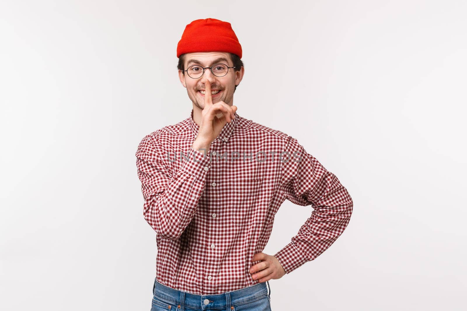 Waist-up portrait of cute happy and kind young bearded man in red beanie prepare surprise, hush at camera with satisfied grin, hold index finger over lips, ask keep secret, be quiet.