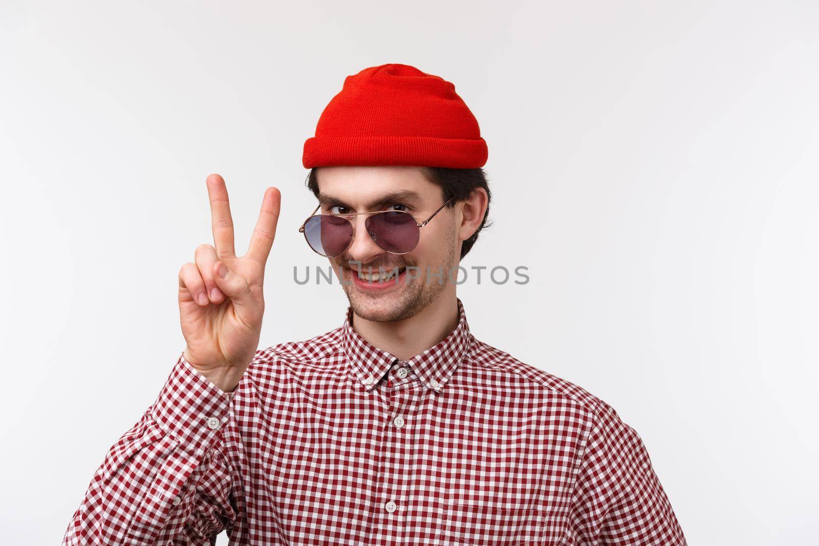 Close-up portrait funny and outgoing hipster man in red beanie, sunglasses and check shirt, getting ready summer vacation, showing peace gesture and smiling, stand white background.