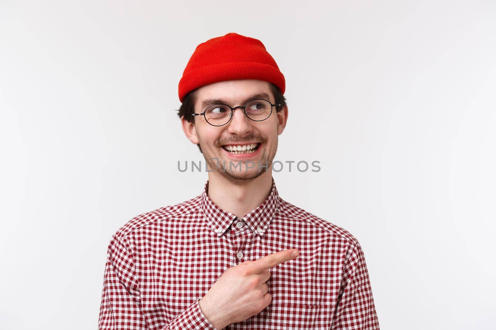 Sly handsome bearded young man in glasses and red beanie, smiling hideous having interesting idea or creative plan, pointing finger upper right corner, standing white background. Copy space