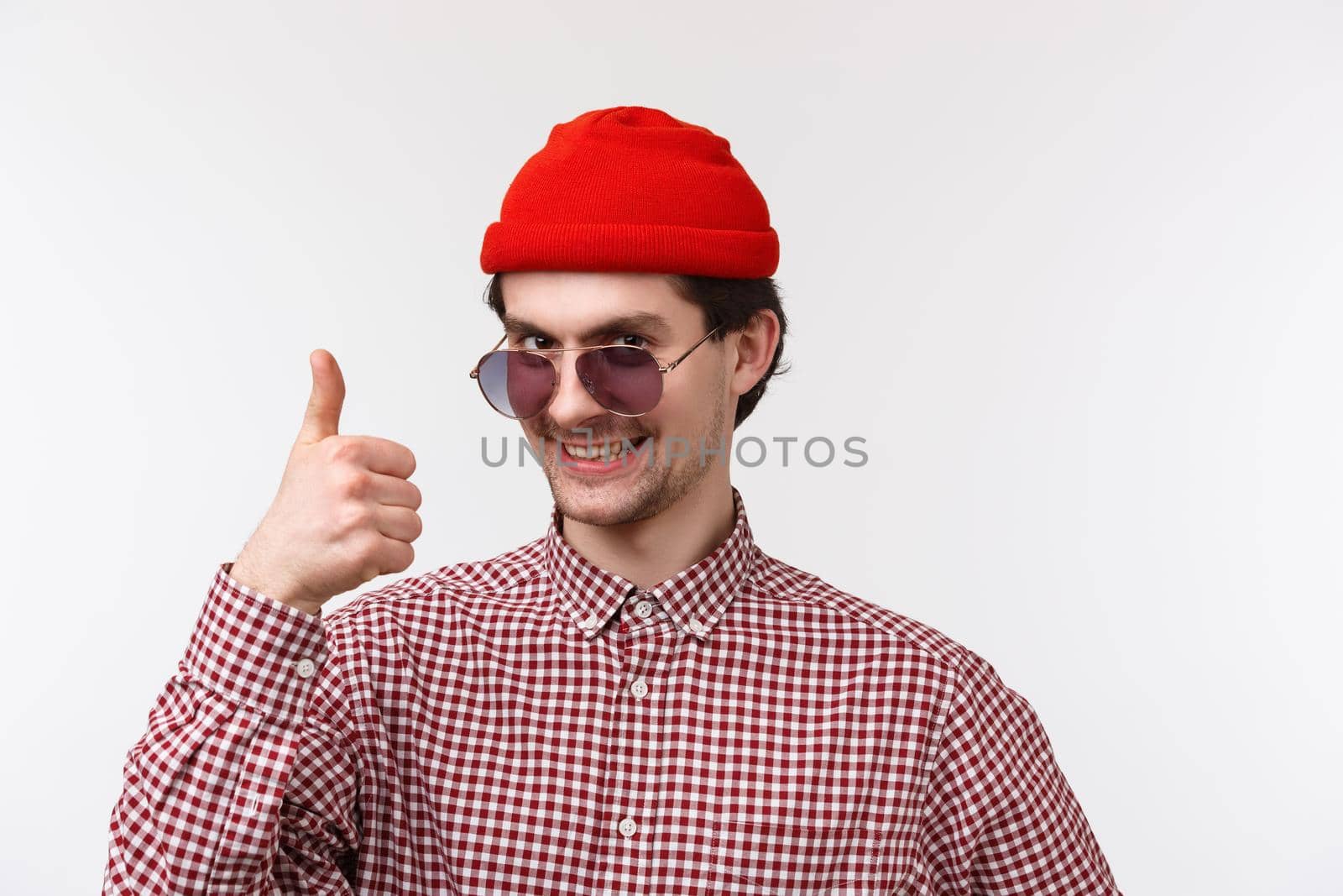Close-up portrait of satisfied handsome and funny young bearded man in checked shirt, hipster red beanie and sunglasses, show thumb-up in like, approval, agree with friend, leave positive comment by Benzoix