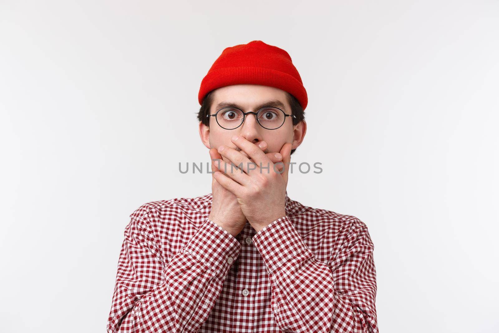Close-up portrait of shocked young man heard something unbelievable and shook, close mouth with hands stare at camera as if gossiping about concerning thing, standing white background.