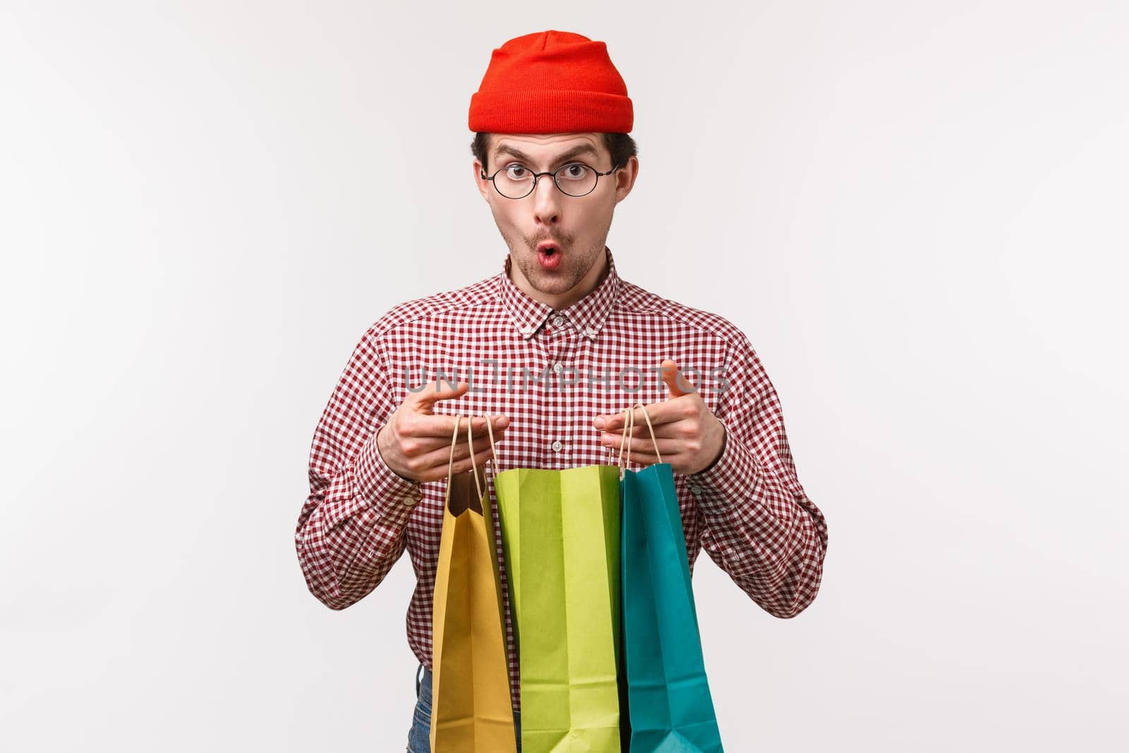 Waist-up portrait excited and surprised handsome young man with moustache, wear glasses and beanie, looking amazed camera say wow as open shopping bags with gifts, stand white background.