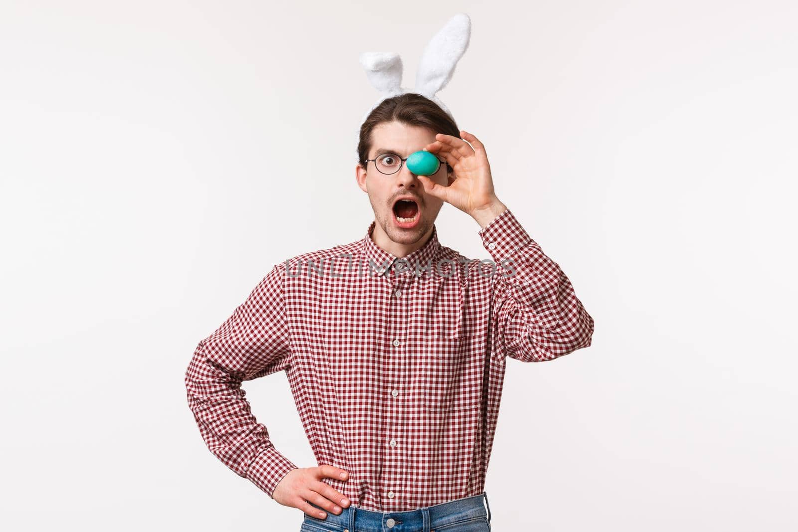 Traditions, religious holidays, celebration concept. Astounded charismatic young bearded man in glasses with cute fake rabbit ears, open mouth surprised, holding painted Easter egg over eye.