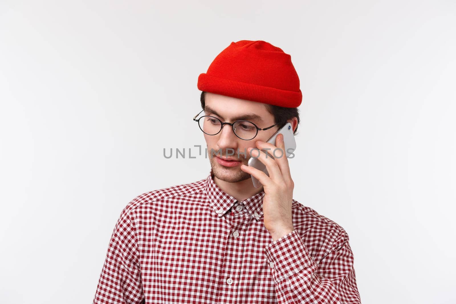 Close-up portrait of serious-looking young man in red beanie and glasses calling someone on mobile phone, having conversation, look sideways as making order or confirm reservation, white background by Benzoix