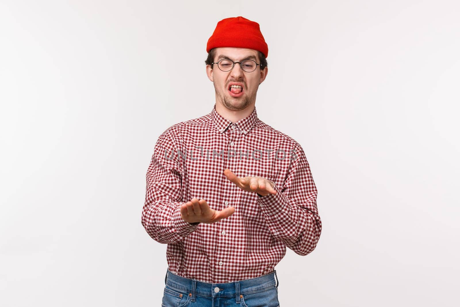 Portrait funny relucant young man in red beanie and glasses, shaking hands in rejection, stick tongue and grimacing disgusted, express disapproval and dislike or awful ugly thing, white background.