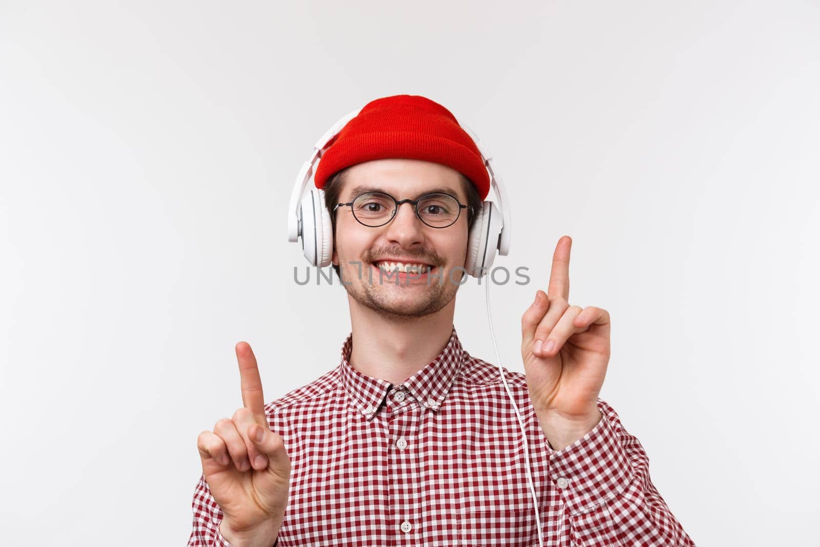 Music, technology and people concept. Close-up portrait of cheerful caucasian man with moustache in glasses and red beanie, dancing moving fingers to rhythm, wear headphones, white background.