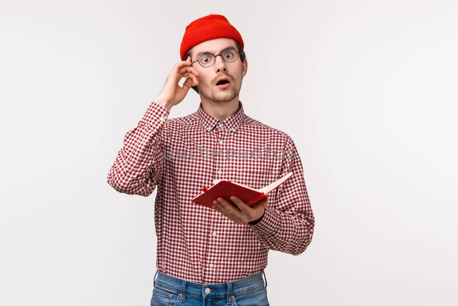Waist-up portrait geeky and creative guy in red beanie, glasses have stunning great idea, touch temple and look inspired while taking notes in notebook, creating equation, stand white background by Benzoix