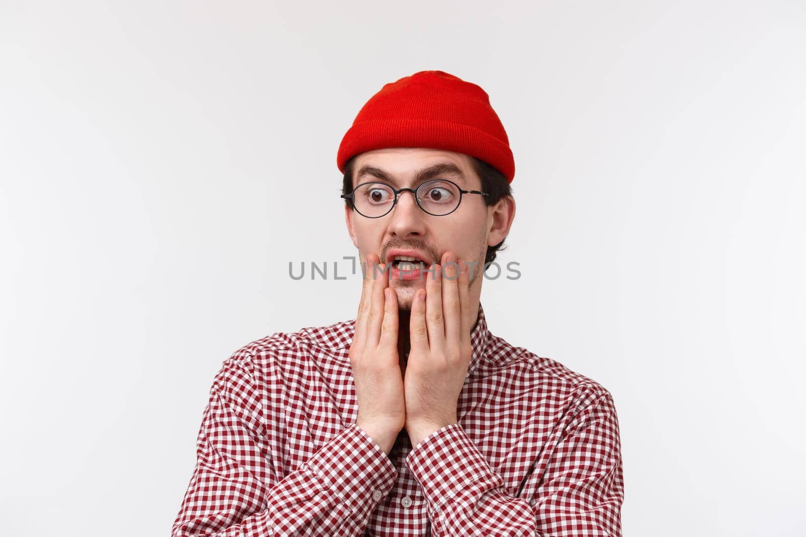 Oh gosh omg. Surprised and speechless hipster guy with beard in red beanie, glasses, gasping staring at something shocking gross, look left spot terrible thing, standing white background.
