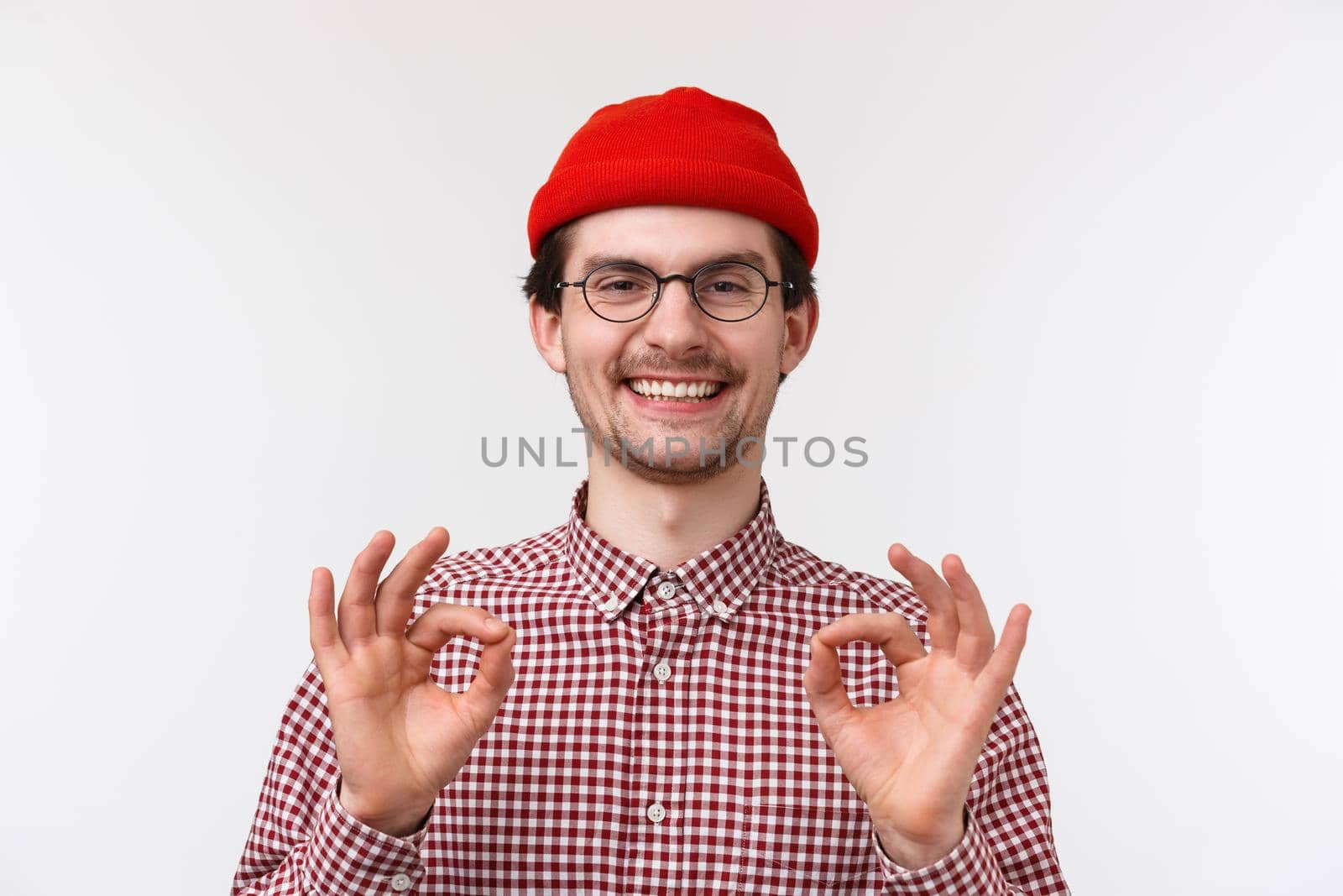 Close-up portrait caucasian male in glasses and red beanie, showing okay gesture rate good movie, being fully satisfied, guarantee all be perfect, standing pleased standing white background.