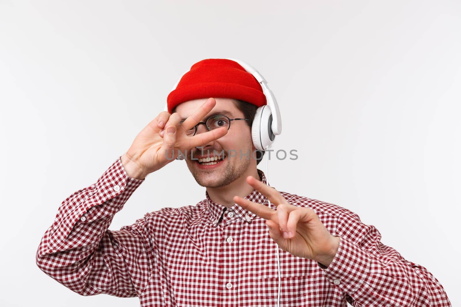 Music, technology and people concept. Close-up portrait happy cheerful young man with moustache and beard, wear hipster red beanie, listening music in headphones and show peace signs.