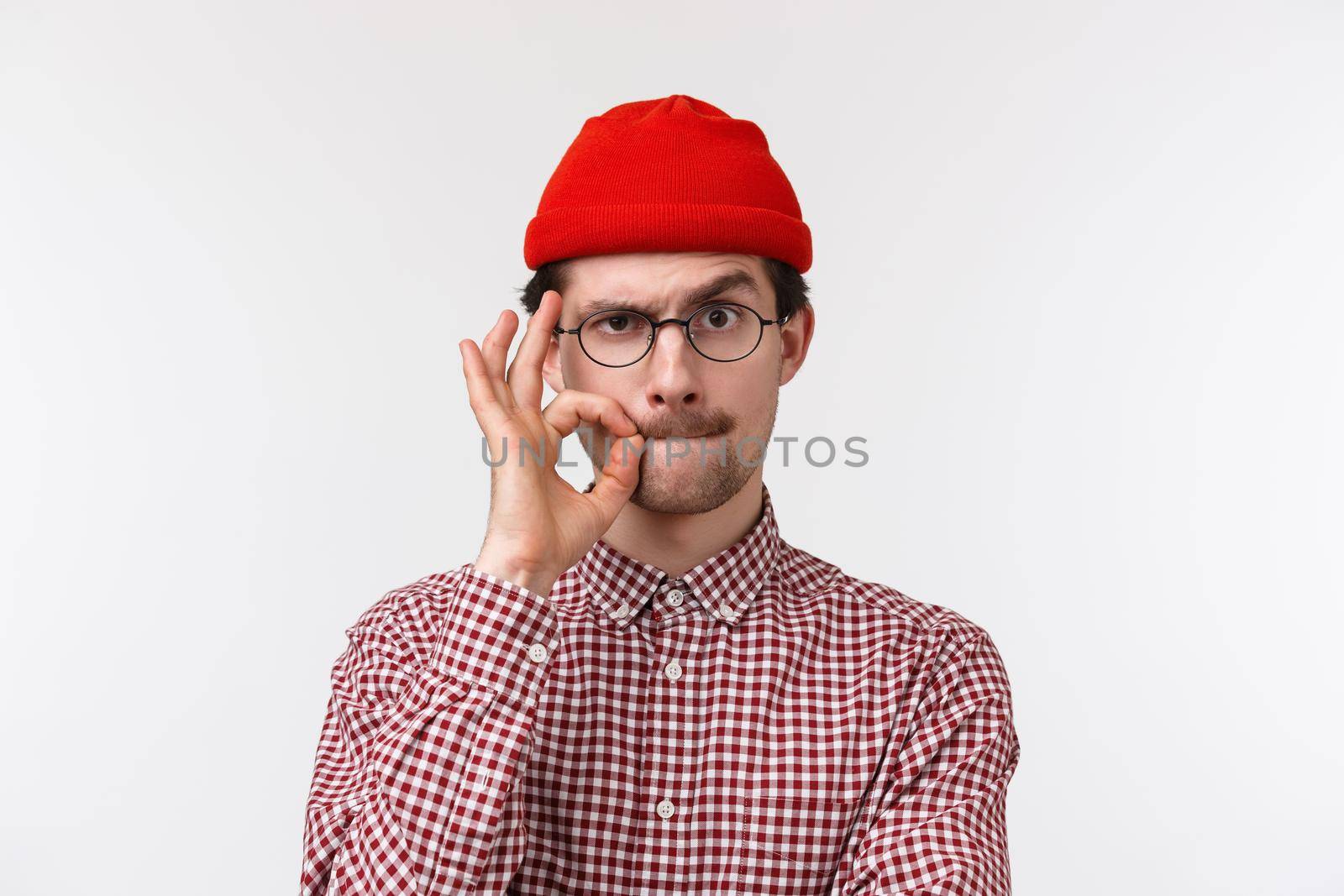 Close-up portrait of serious young caucasian guy with moustache, wear glasses and beanie, seal lips with finger gesture as if hiding secret, promise not speak about it, stand white background.