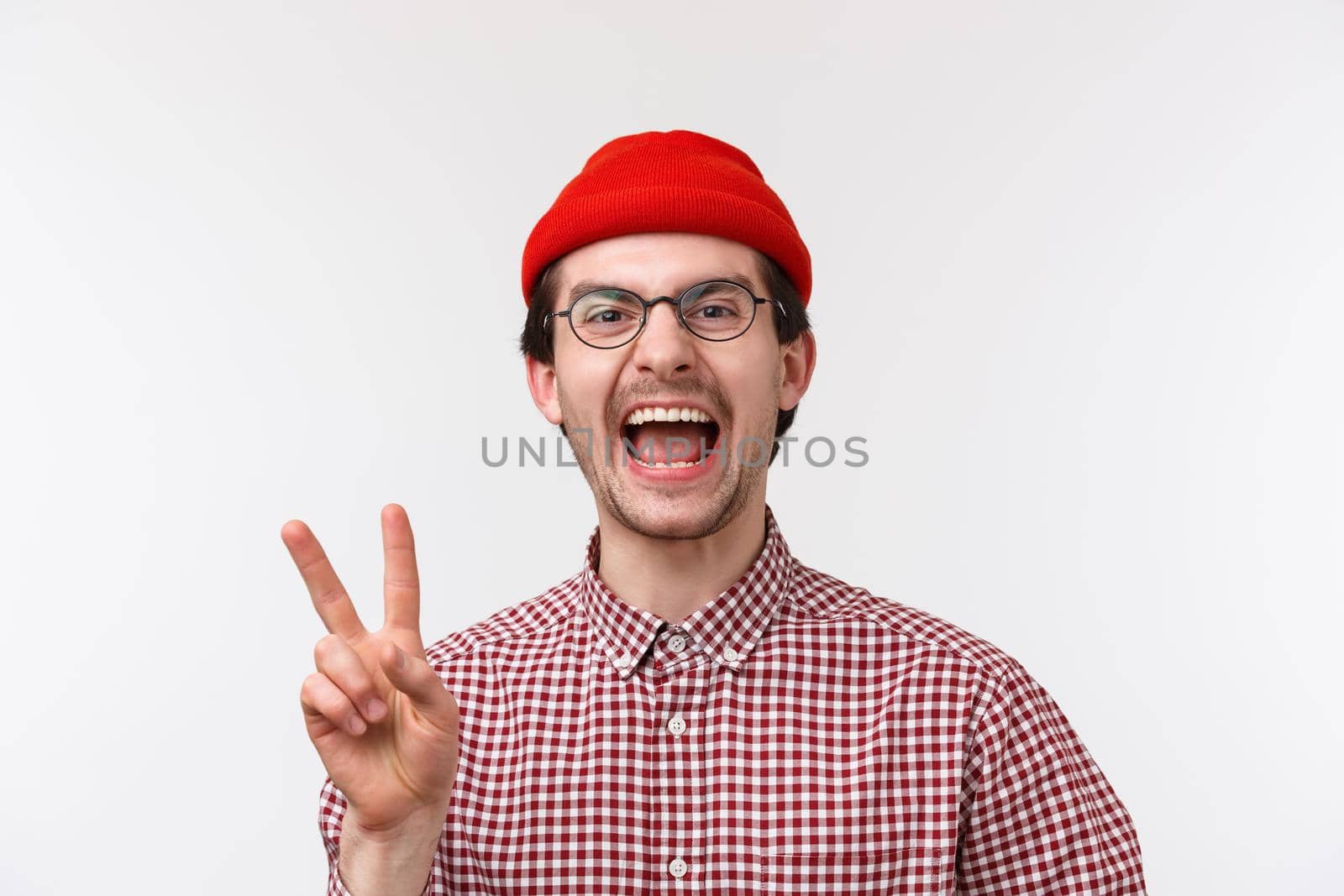 Close-up portrait cheerful and funny caucasian bearded male in glasses and red beanie, laughing and showing peace gesture looking camera happy, standing white background by Benzoix