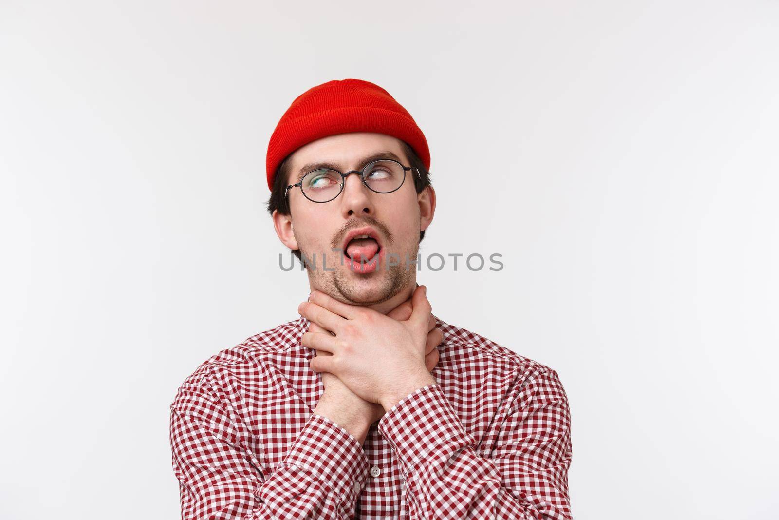 Close-up portrait of funny bearded guy chocking himself stick tongue as if cant breath, roll eyes pretend dying from boredom or express his distress, standing in red beanie and glasses by Benzoix