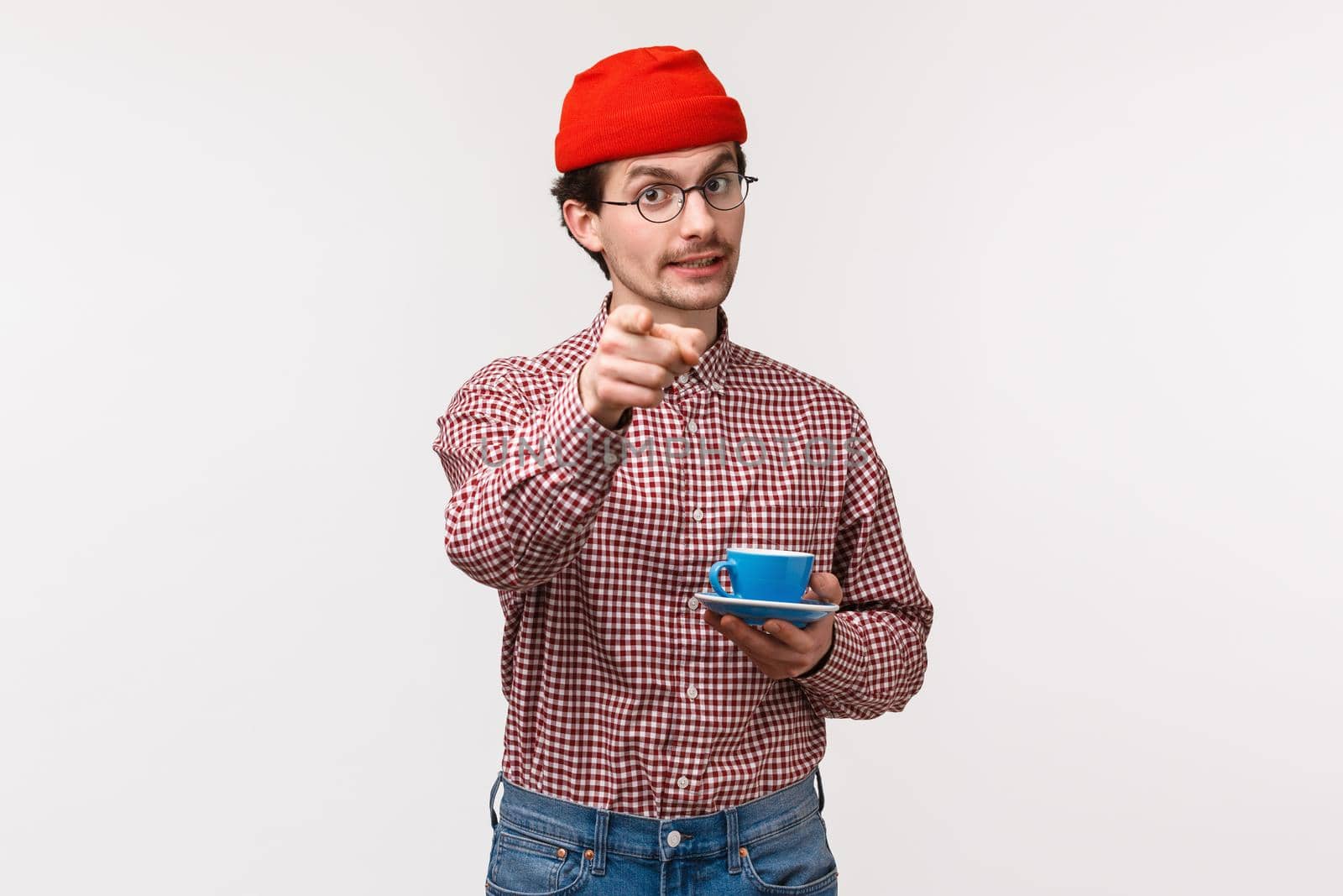 Waist-up portrait of handsome cheerful caucasian bearded man in glasses and red beanie, holding cup of coffee or tea and pointing at camera as telling to try this awesome drink yourself by Benzoix