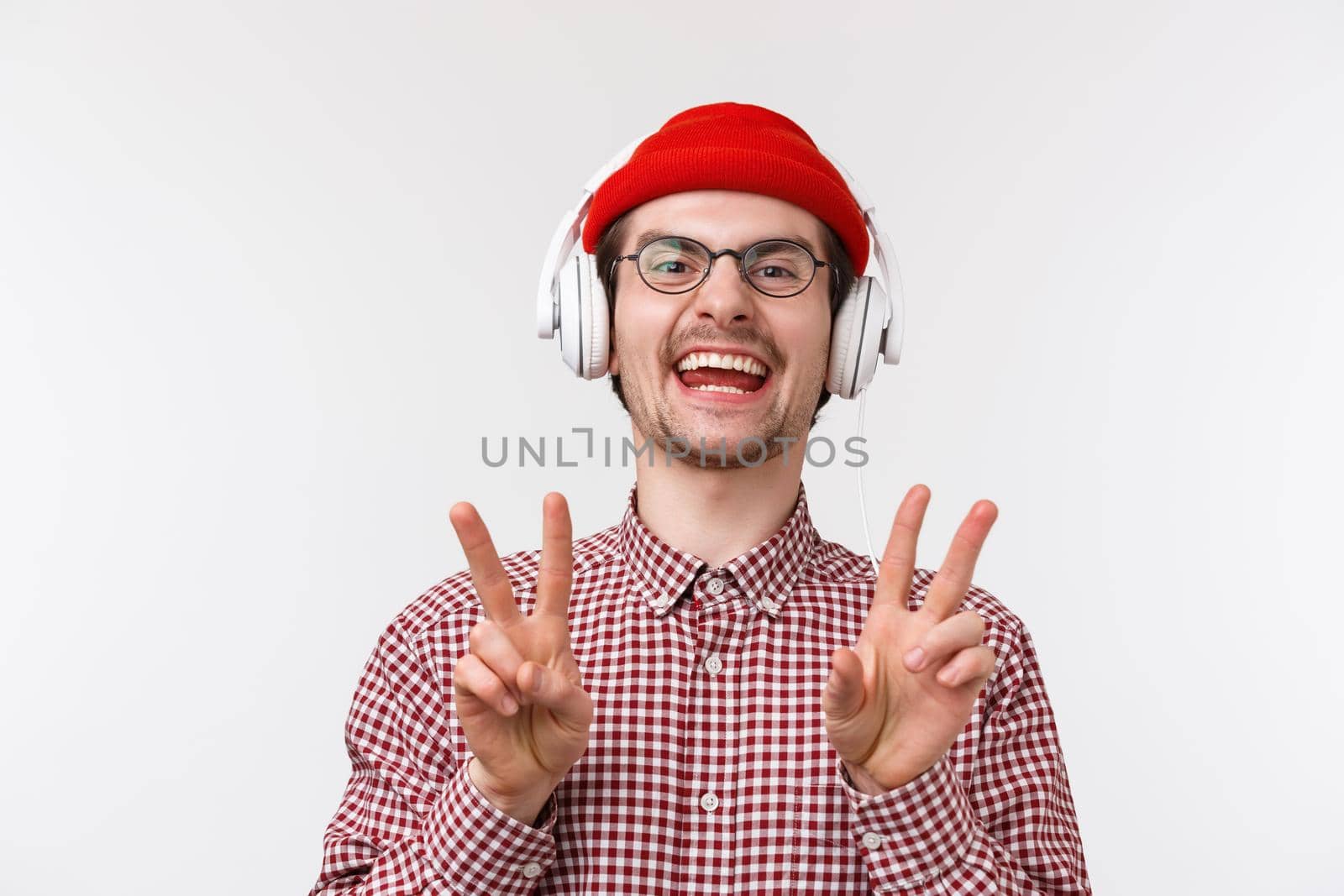 Music, technology and people concept. Positive and happy smiling young man with beard and glasses, wear beanie listen music in headphones, showing peace gesture and laughing, white background by Benzoix