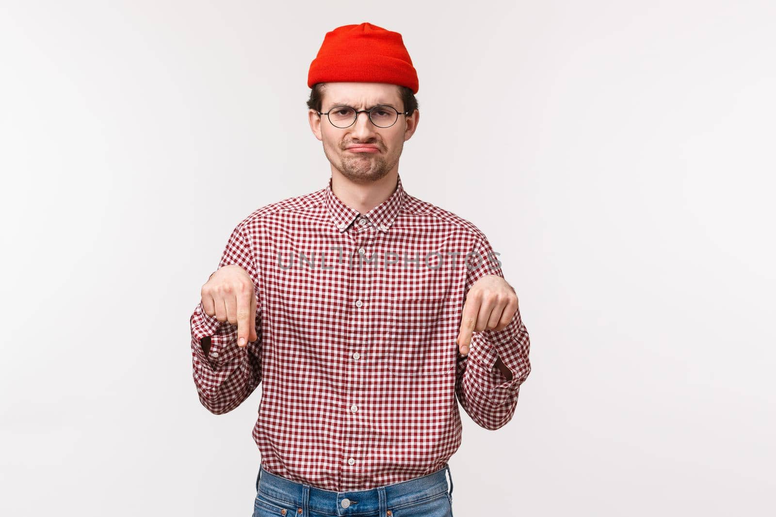 Waist-up portrait funny skeptical and unimpressed bearded man in glasses and red beanie, griamcing disapproval, pointing fingers down uninteresting awful product, stand white background.