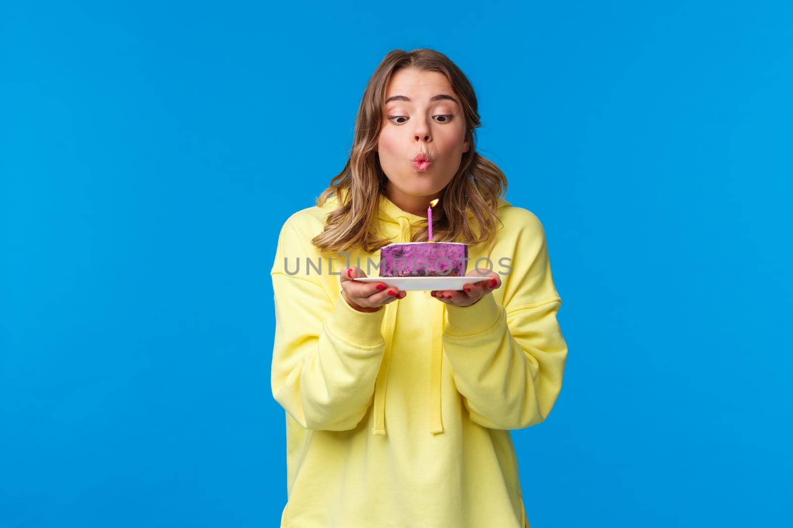 Celebration, party and lifestyle concept. Silly cute european blond woman in yellow hoodie, making wish holding fruit cake blowing out b-day candle, standing blue background at birthday party.