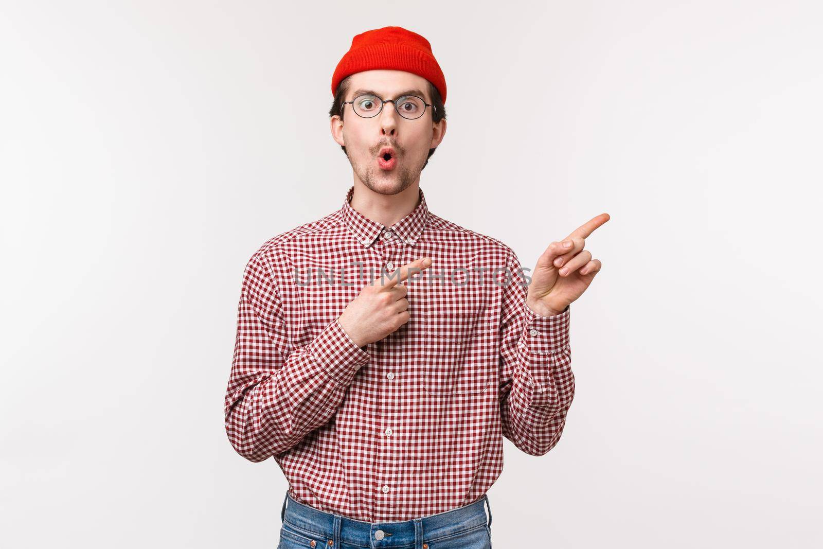 Surprised and curious funny bearded man in red beanie, glasses folding lips from amazement and interest asking question about cool new product in stock, pointing upper right corner by Benzoix