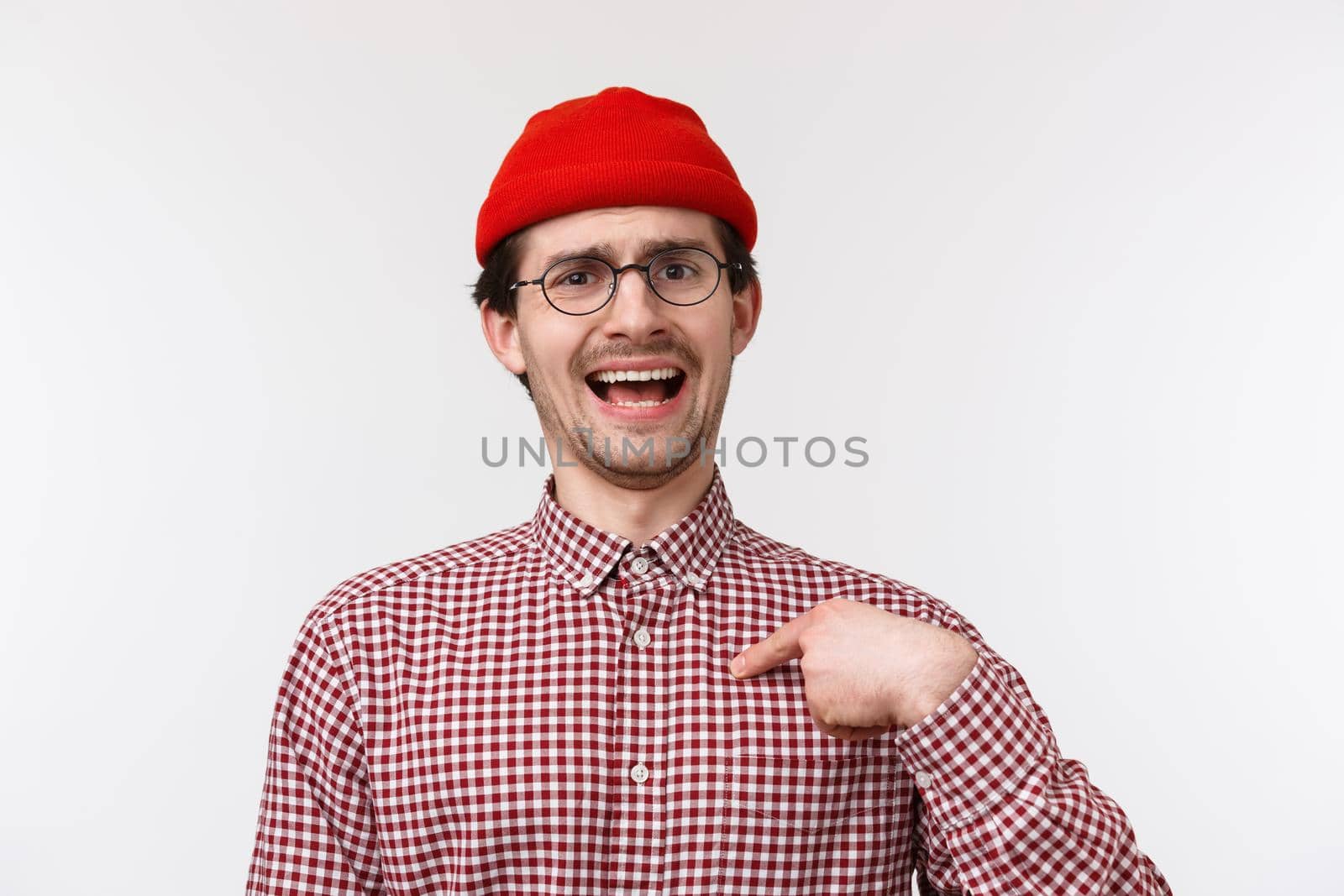 Close-up portrait boastful funny bearded young man in red beanie and glasses, pointing at himself as talking own achievement, bragging personal goals, standing white background.