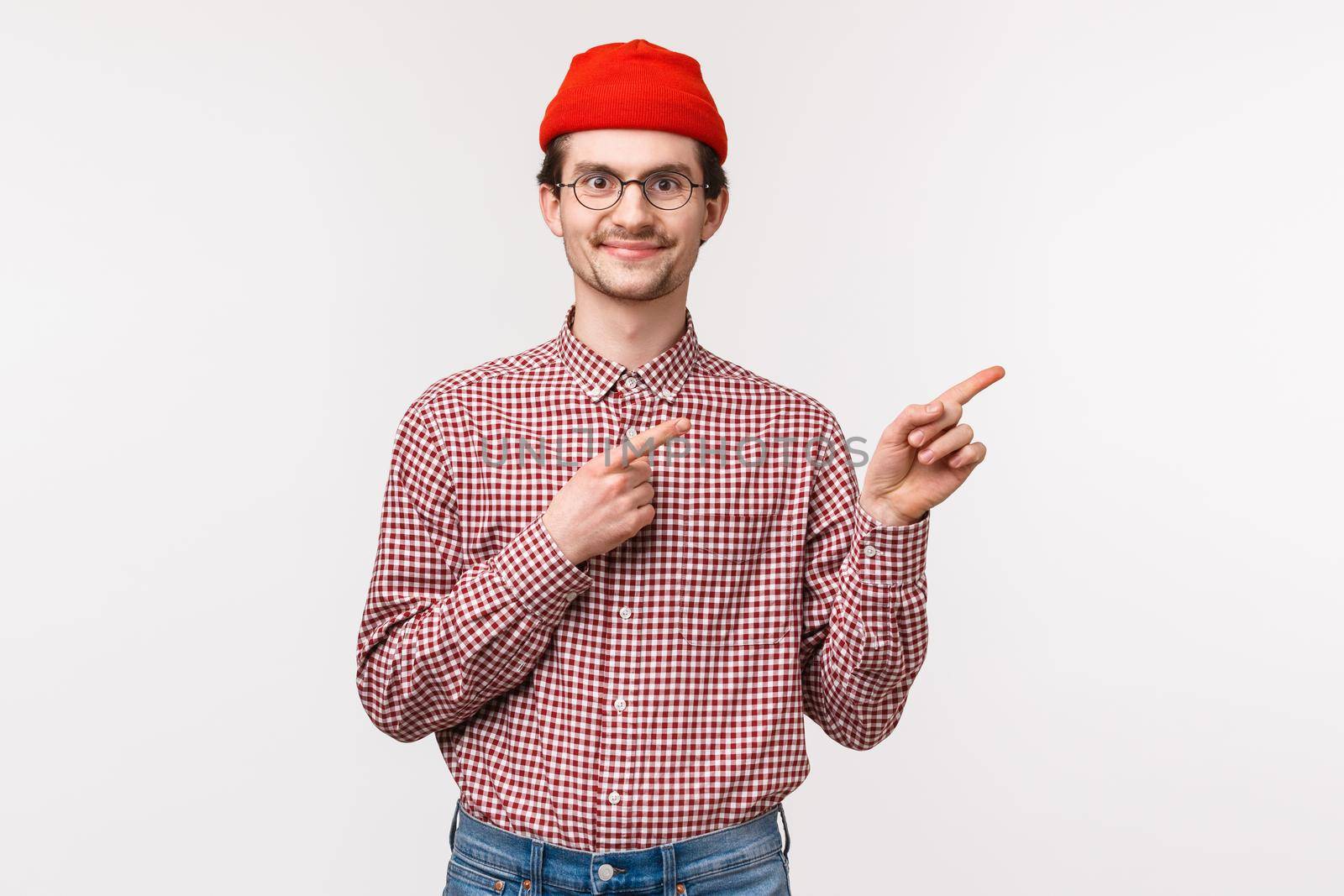 Waist-up portrait of satisfied happy smiling young hipster guy in red beanie, glasses pointing fingers right top advertisement with pleased expression, found excellent product or company service by Benzoix