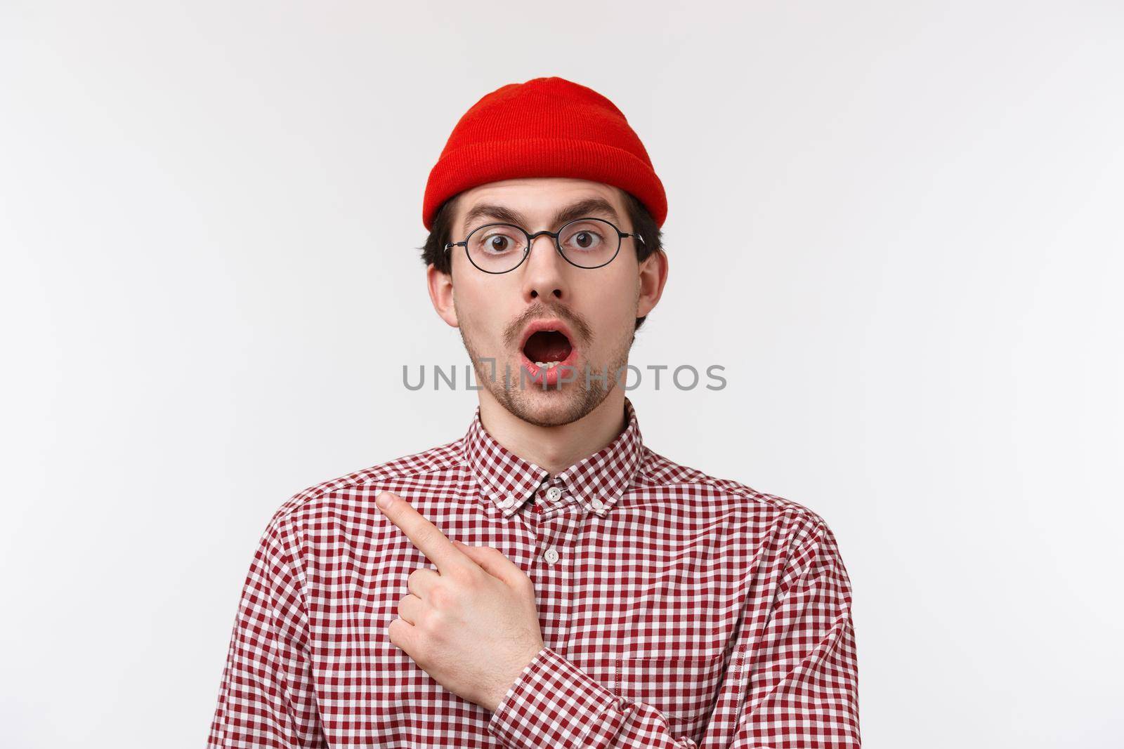 Surprised and impressed handsome bearded hipster man in glasses and red beanie, open mouth and drop jaw gasping stunned, saw something shocking, pointing upper left corner, white background by Benzoix