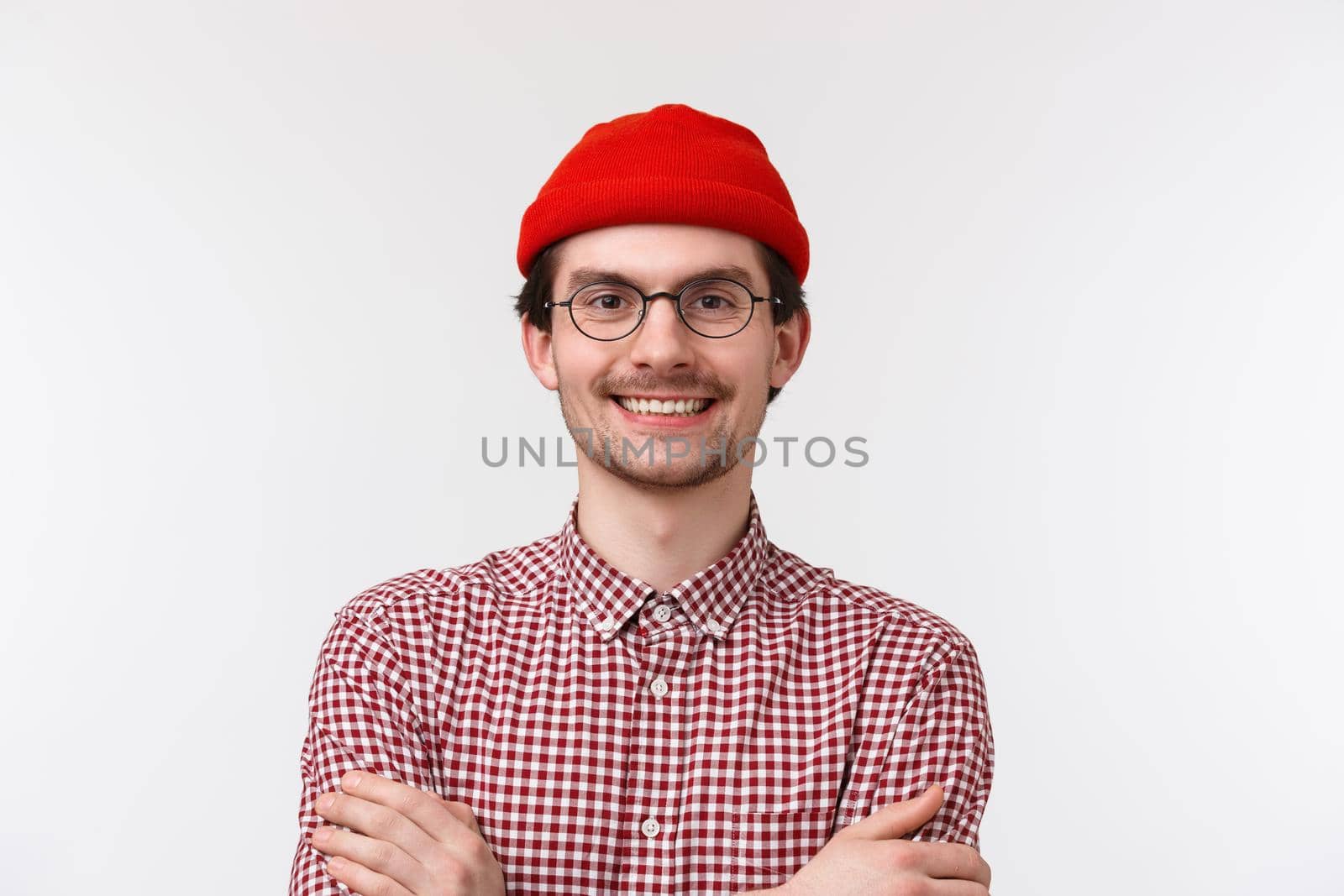 Close-up portrait of confident cheerful young man expressing himself with bright outfit, wear hipster red beanie and checked shirt, cross hands on chest like pro, smiling delighted, white background.